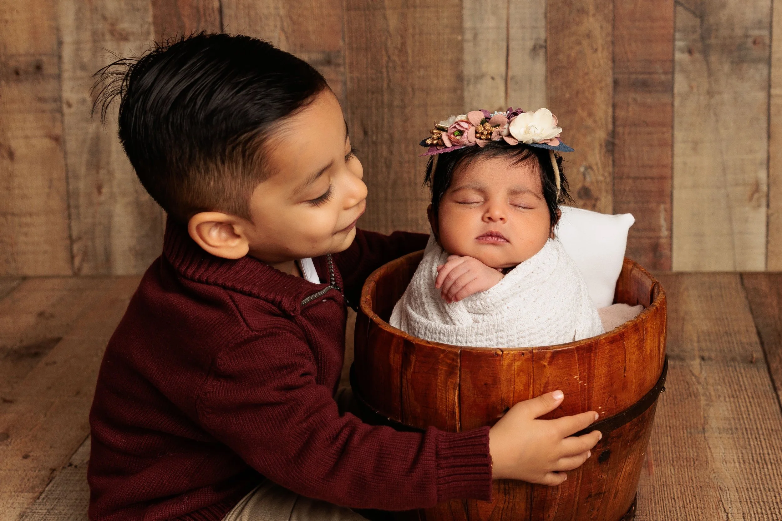 A young boy with dark hair and a burgundy sweater holds a sleeping baby in a wooden basket. The baby is wrapped in a white blanket and wears a floral headband. They are posed against a rustic wooden background.