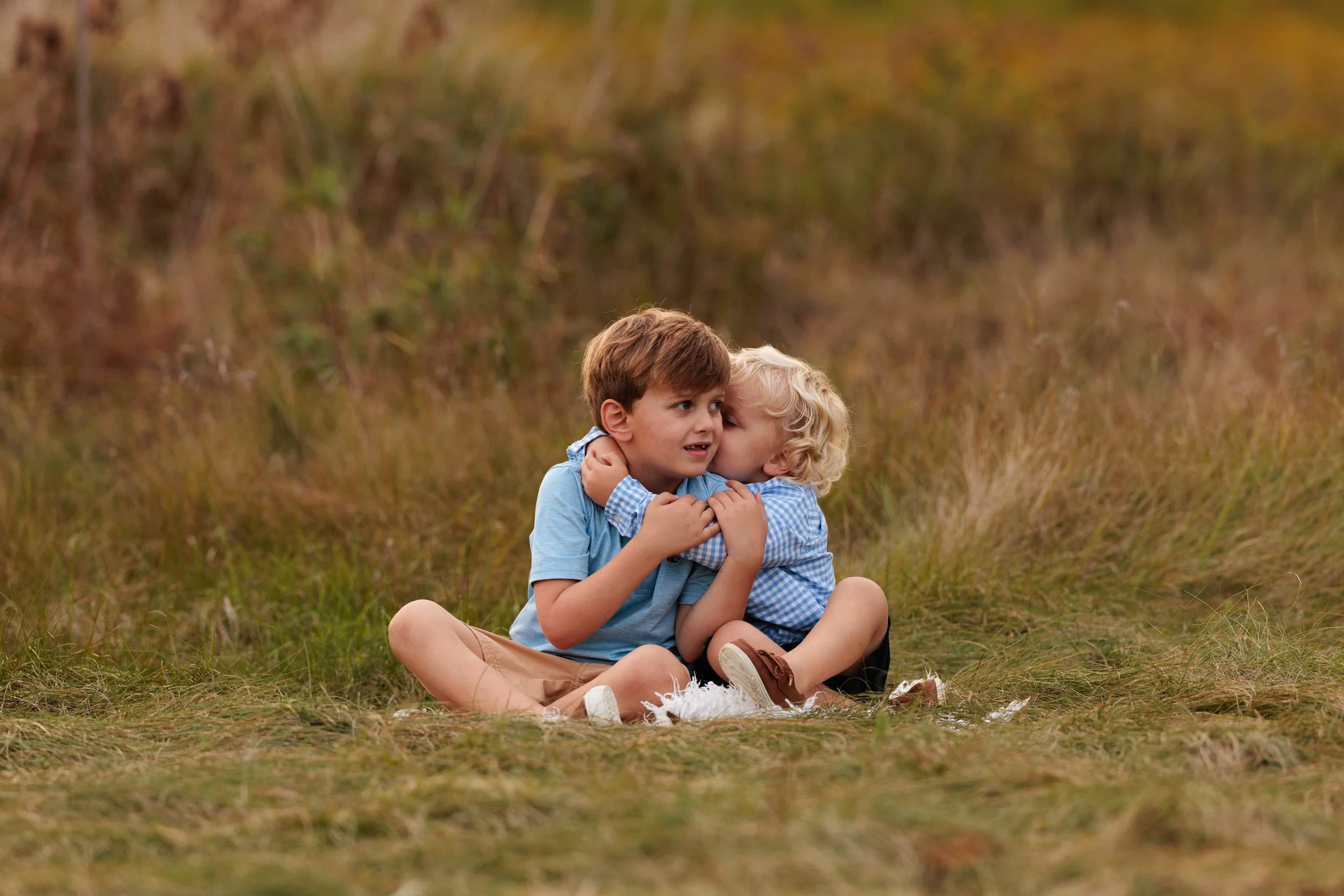 Two young boys sitting on grass in a field embracing, one kissing the other on the cheek.