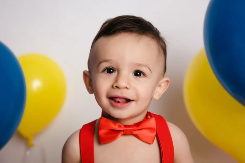 A smiling young boy with short dark hair, wearing a red bow tie and suspenders, surrounded by yellow and blue balloons.