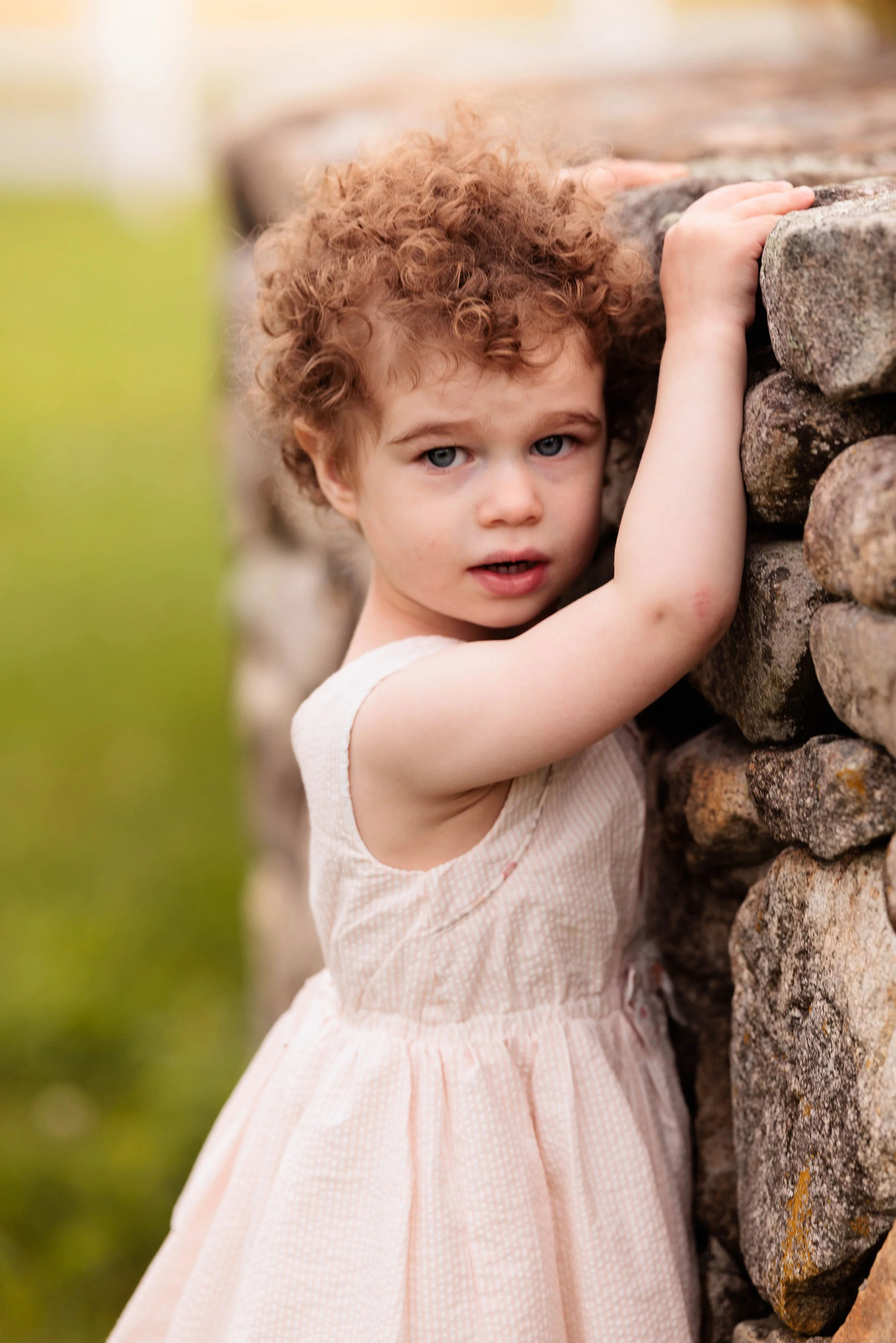 A young girl with curly red hair and blue eyes, wearing a sleeveless light pink dress, holding onto a stone wall outdoors.