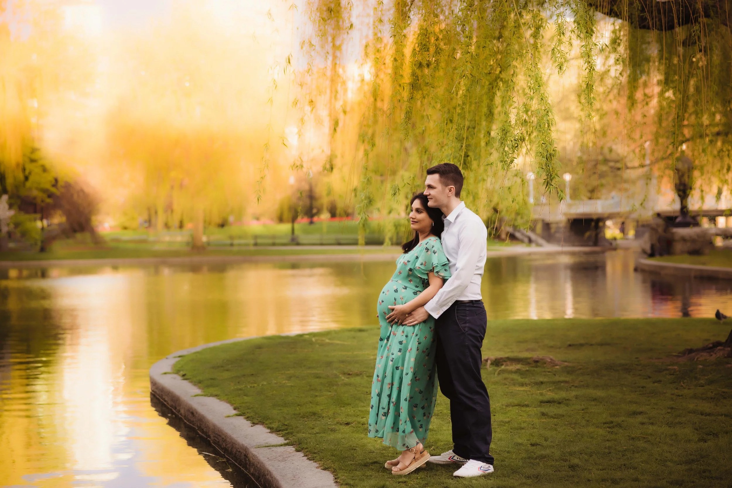 A pregnant woman and a man standing by a lake in a park during sunset, with trees hanging over them and a warm, golden light reflecting on the water.