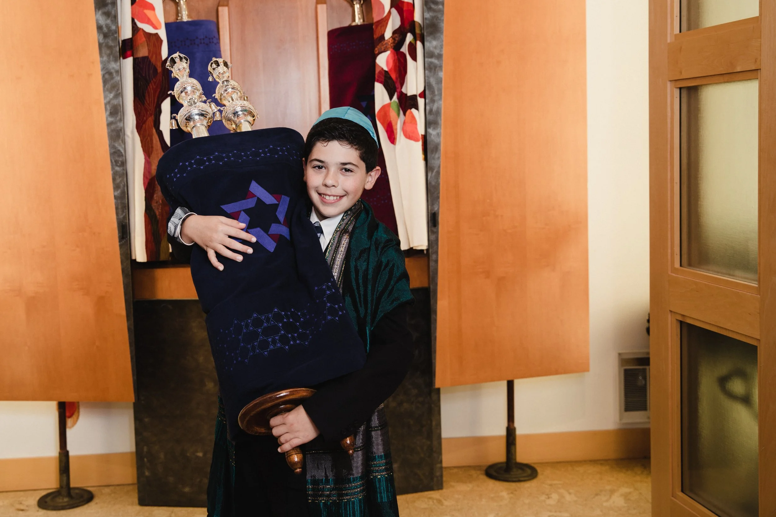 A young boy smiling and holding a large menorah in a room with wooden furniture and patterned curtains.