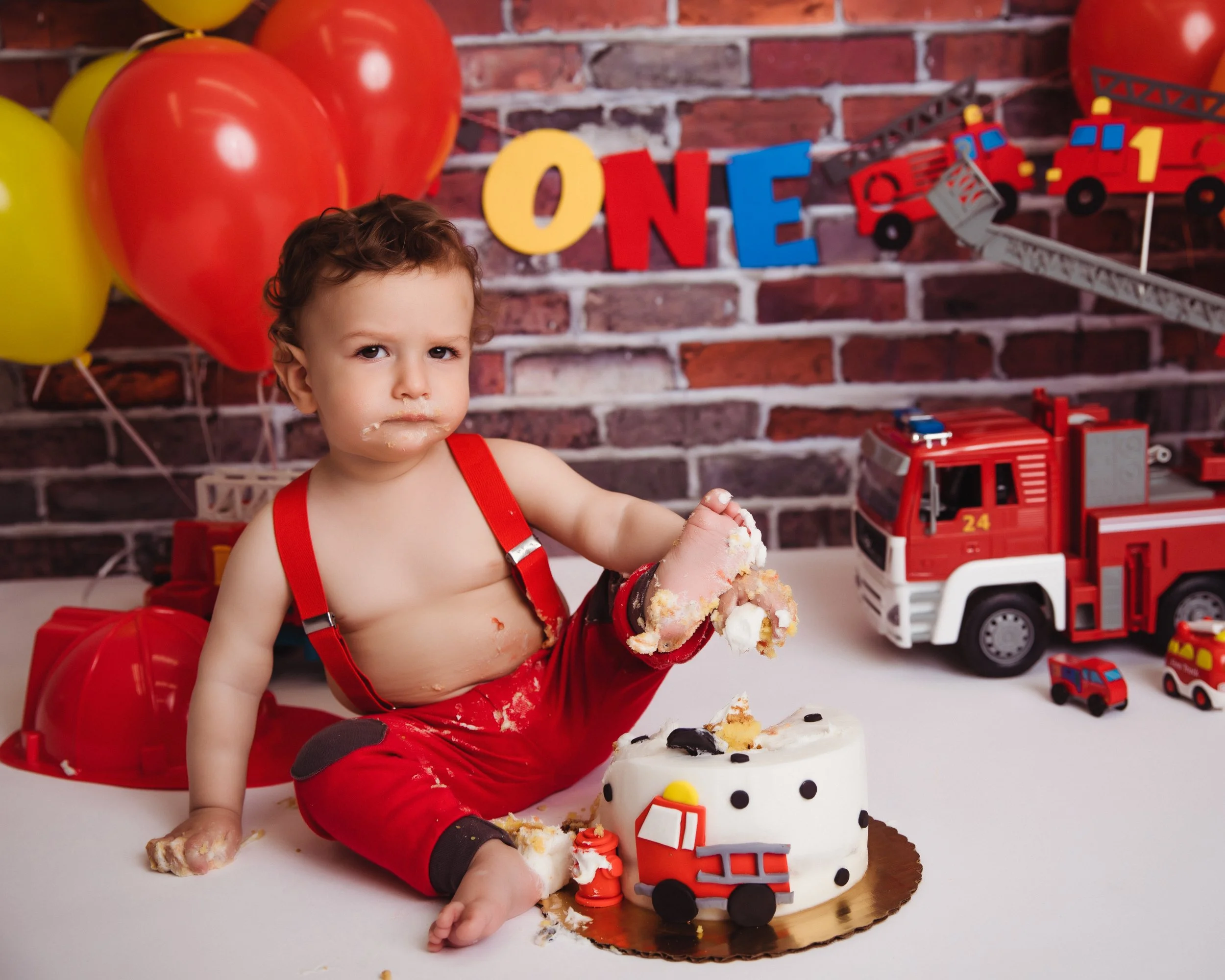 A young boy sitting at a birthday celebration with a fire truck-themed cake, surrounded by toy fire trucks, balloons, and colorful alphabet and fire truck decorations in the background.