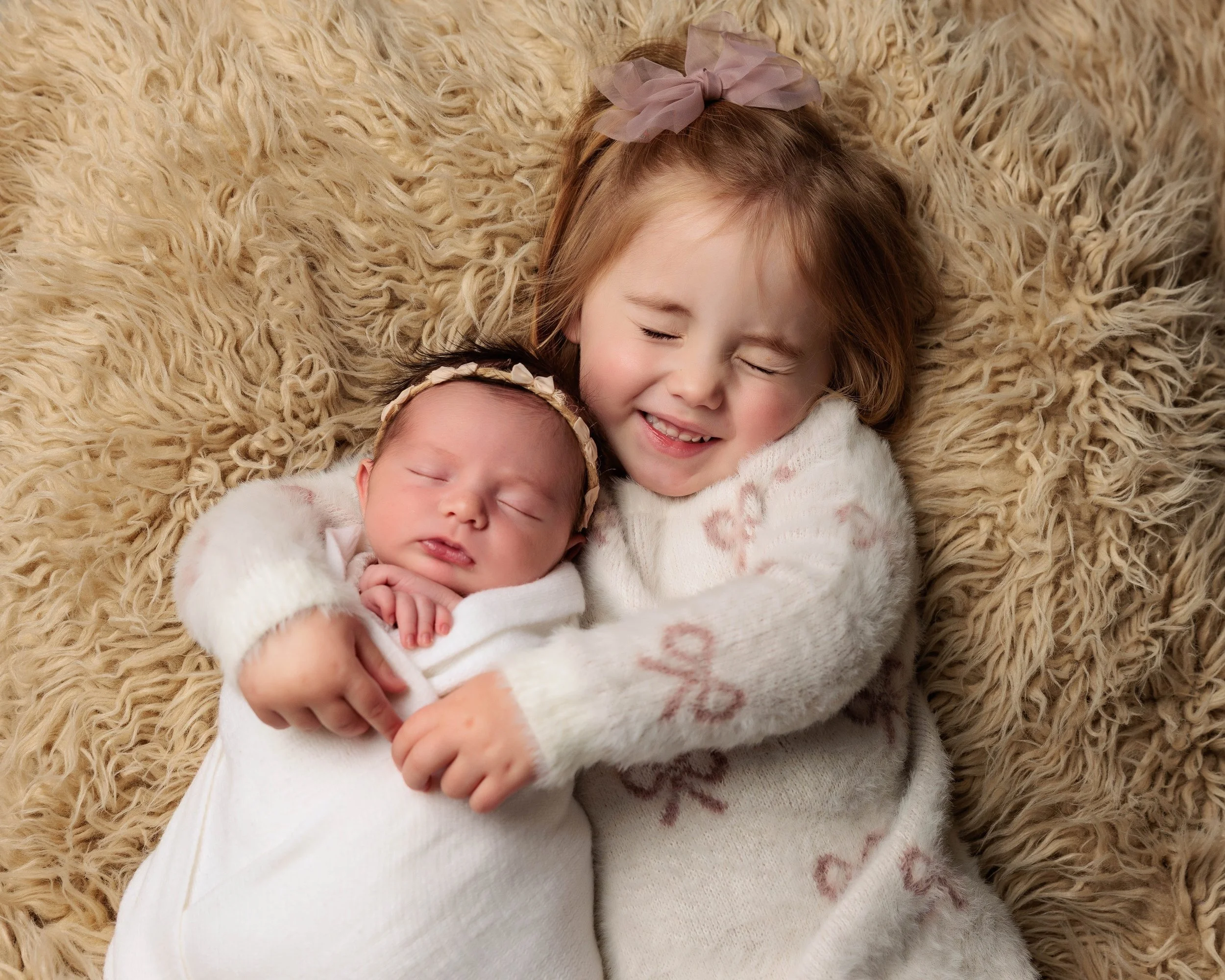 A young girl with a pink bow hugging a sleeping baby girl, both on a plush beige blanket.