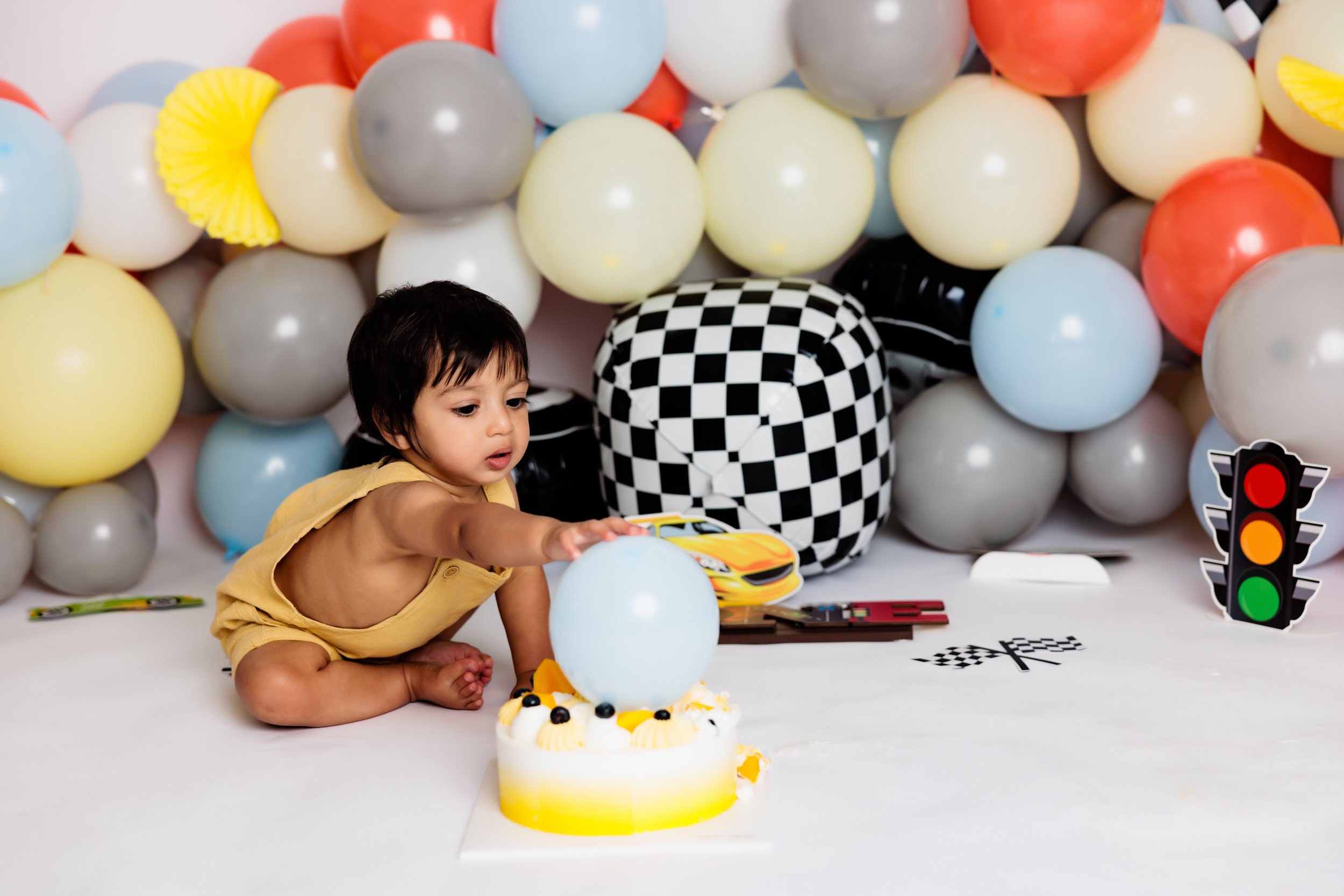 Young child in a yellow outfit sitting on the floor, reaching towards a white and yellow birthday cake with a large blue balloon on top. The background is decorated with colorful balloons and various themed decorations, including a checkered box and 