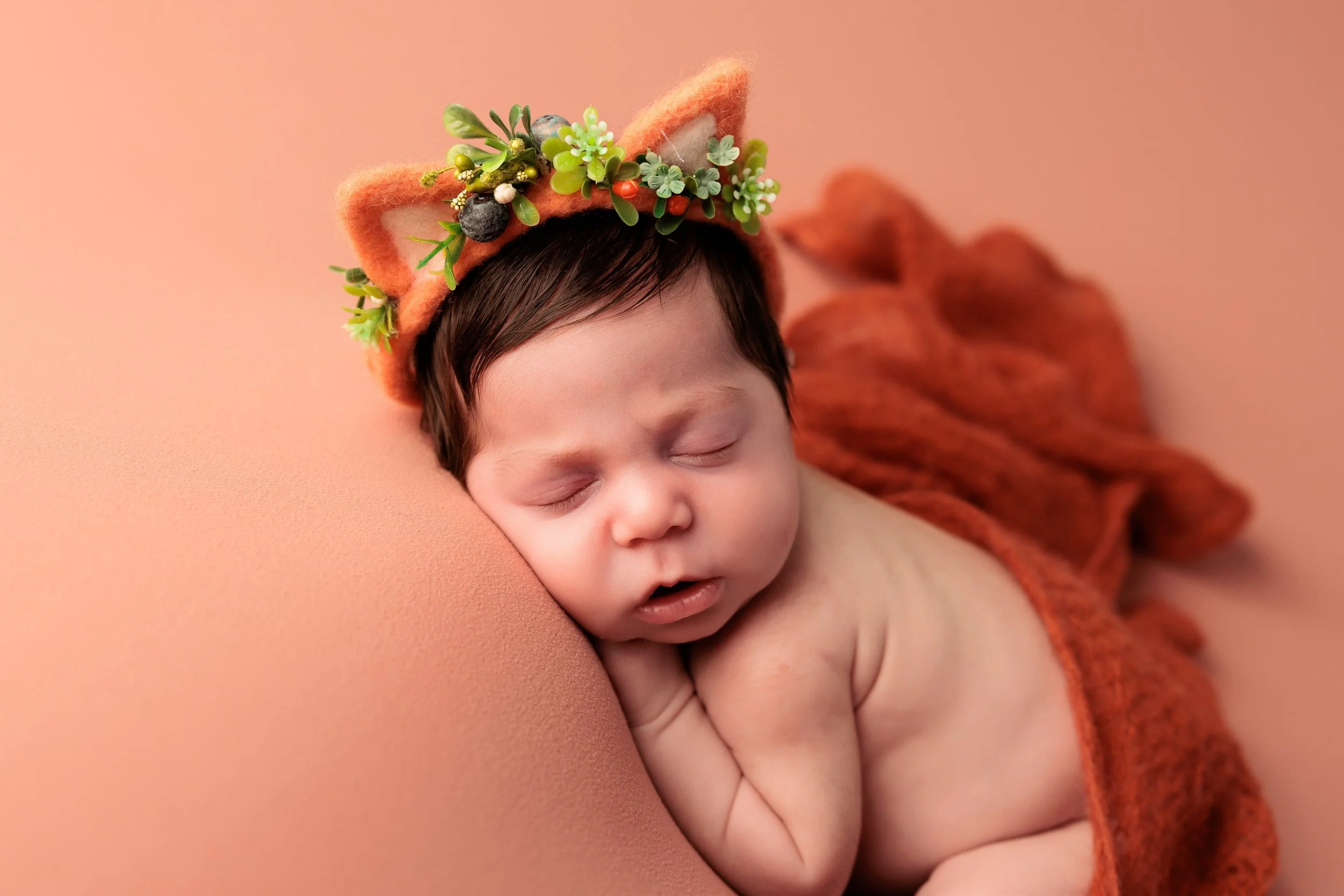 A sleeping baby with a fox-themed headband and a rust-colored blanket on a pink background.