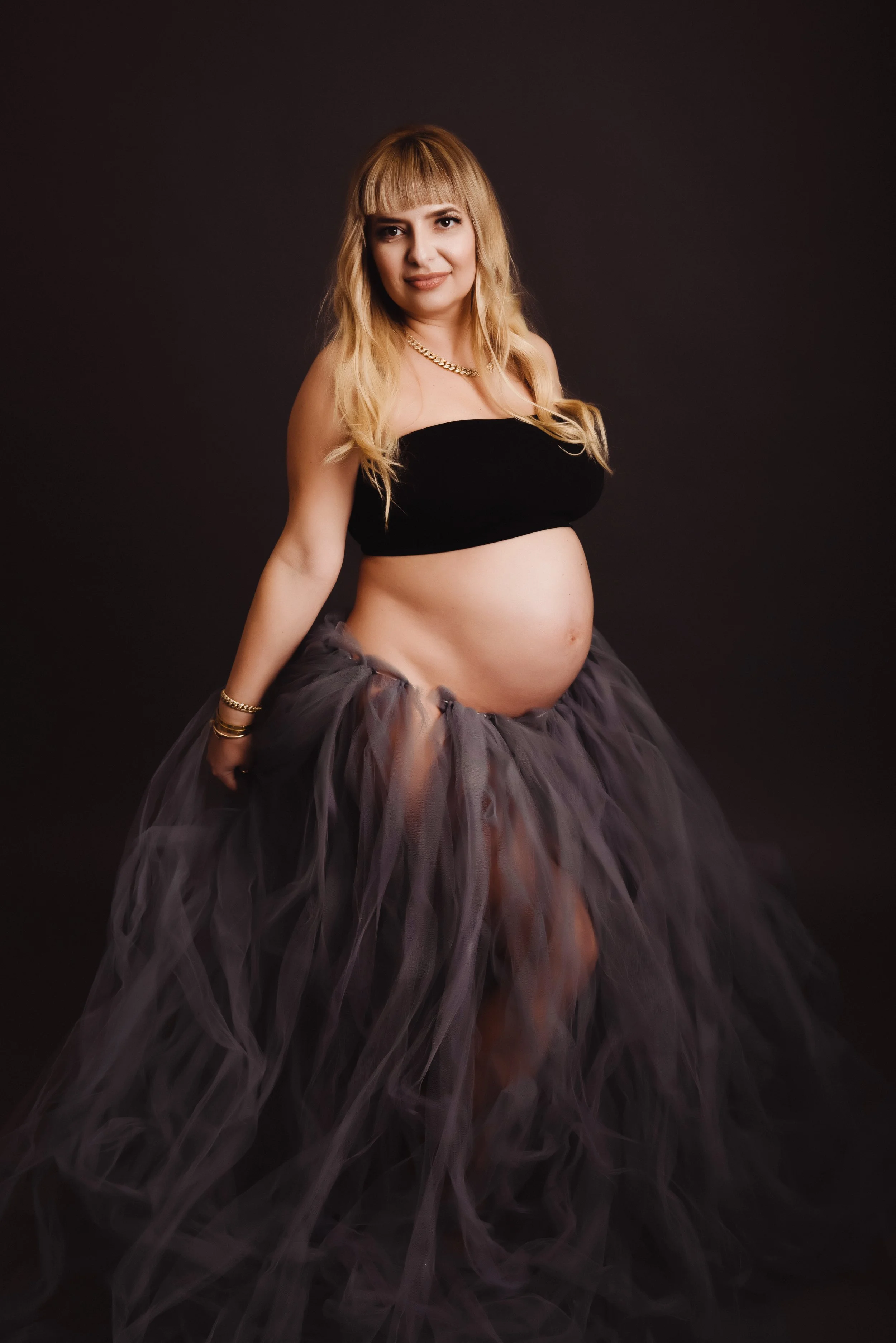 A pregnant woman with blonde hair wearing a black band top and a flowing purple tulle skirt, posing against a black background.