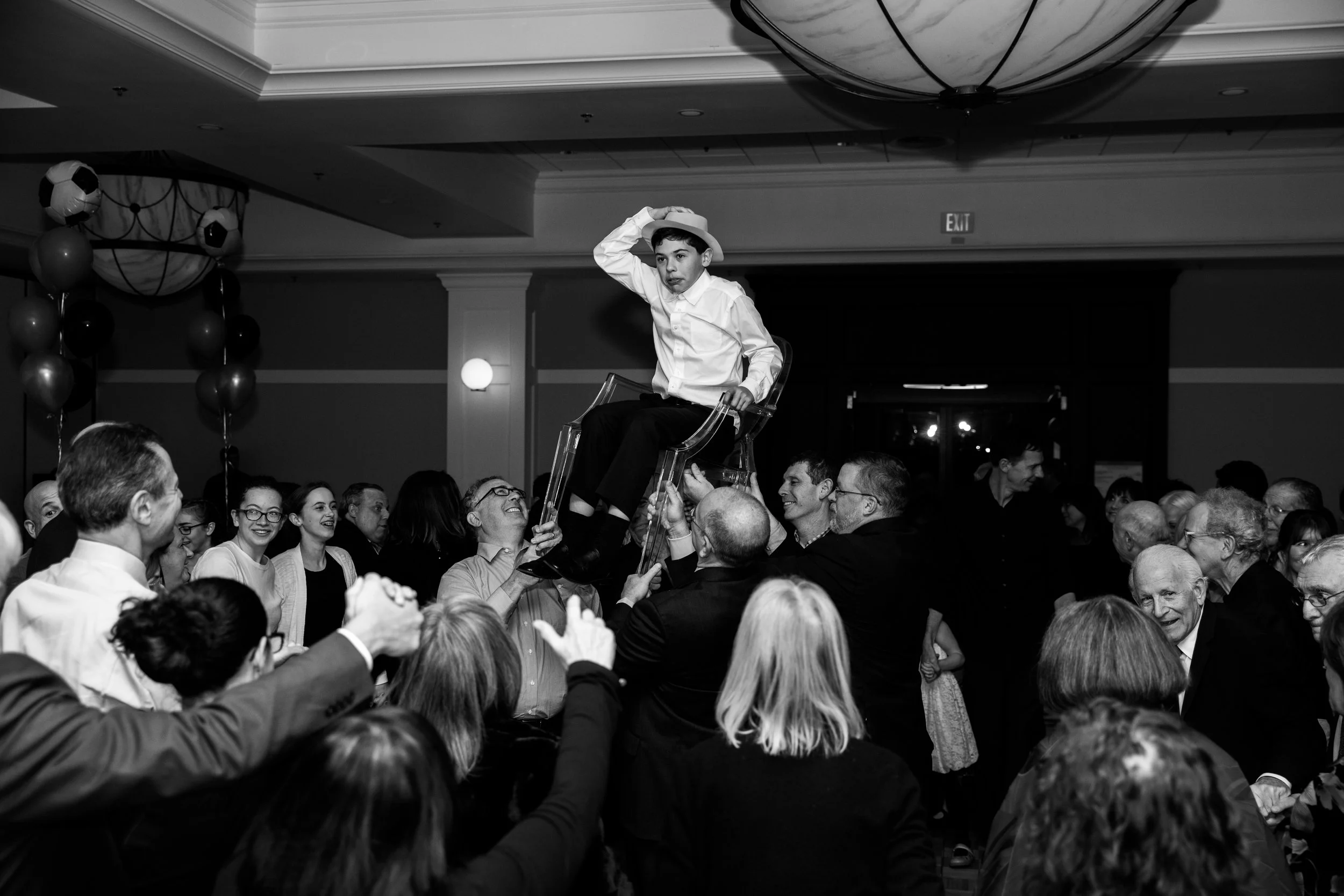 A boy in a white shirt and hat sitting on a transparent chair being lifted by a group of people at a lively event with balloons and atmospheric lighting.