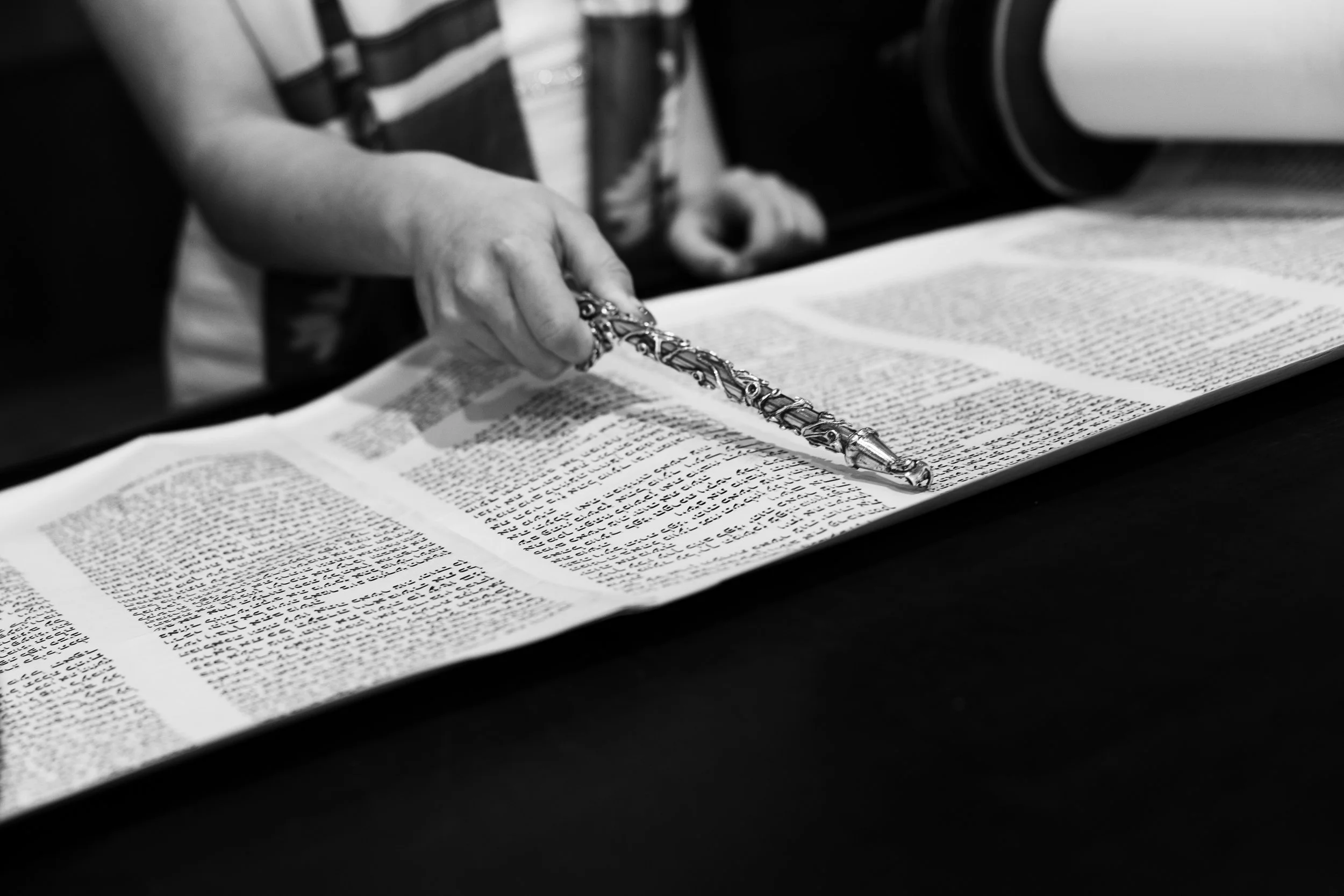 A person using a decorative silver pointer to read a large open book, with a focus on their hand and the pointer in black and white.