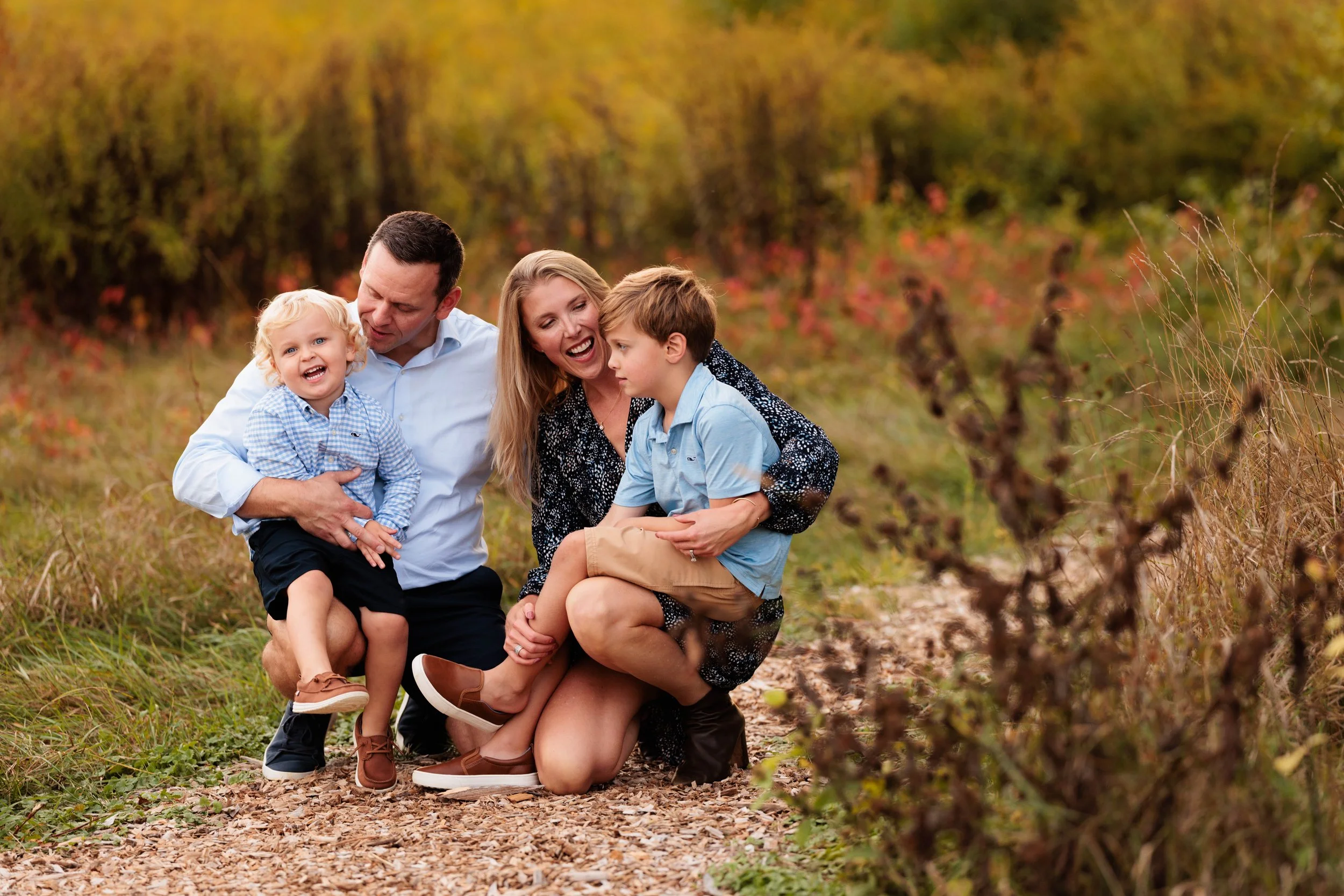 A family of four enjoying time outdoors on a nature trail in autumn with colorful trees in the background, laughing and playing.