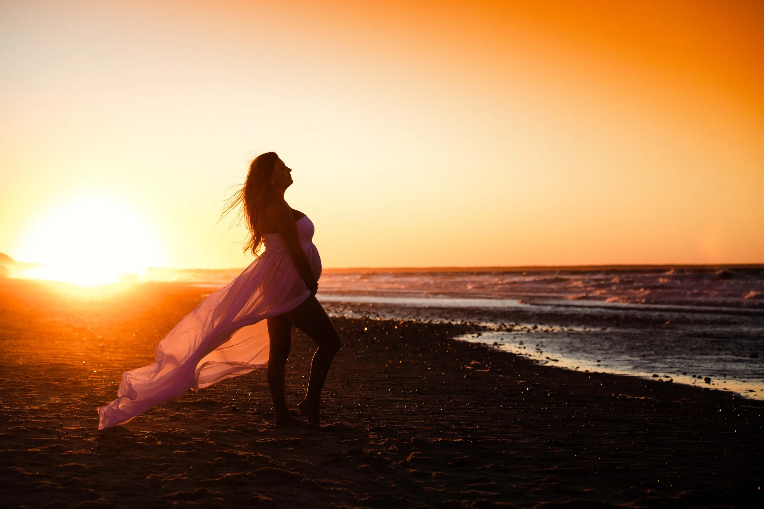 A pregnant woman standing on a beach at sunset, wearing a flowing dress, with her hair blowing in the wind.