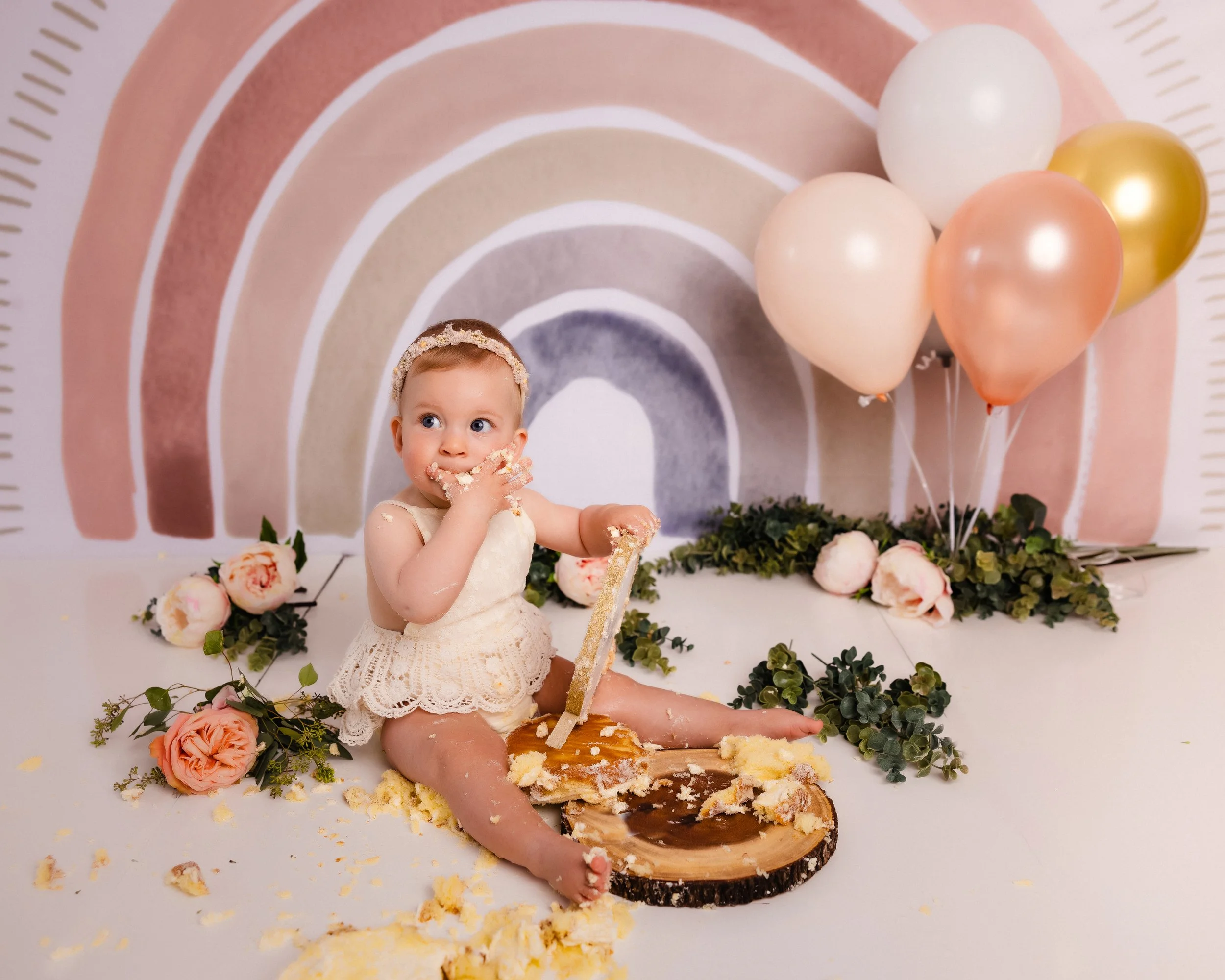 Baby girl sitting on the floor in front of a rainbow-themed backdrop, surrounded by pink and white flowers and balloons, eating cake with a piece of cake on a wooden platter.