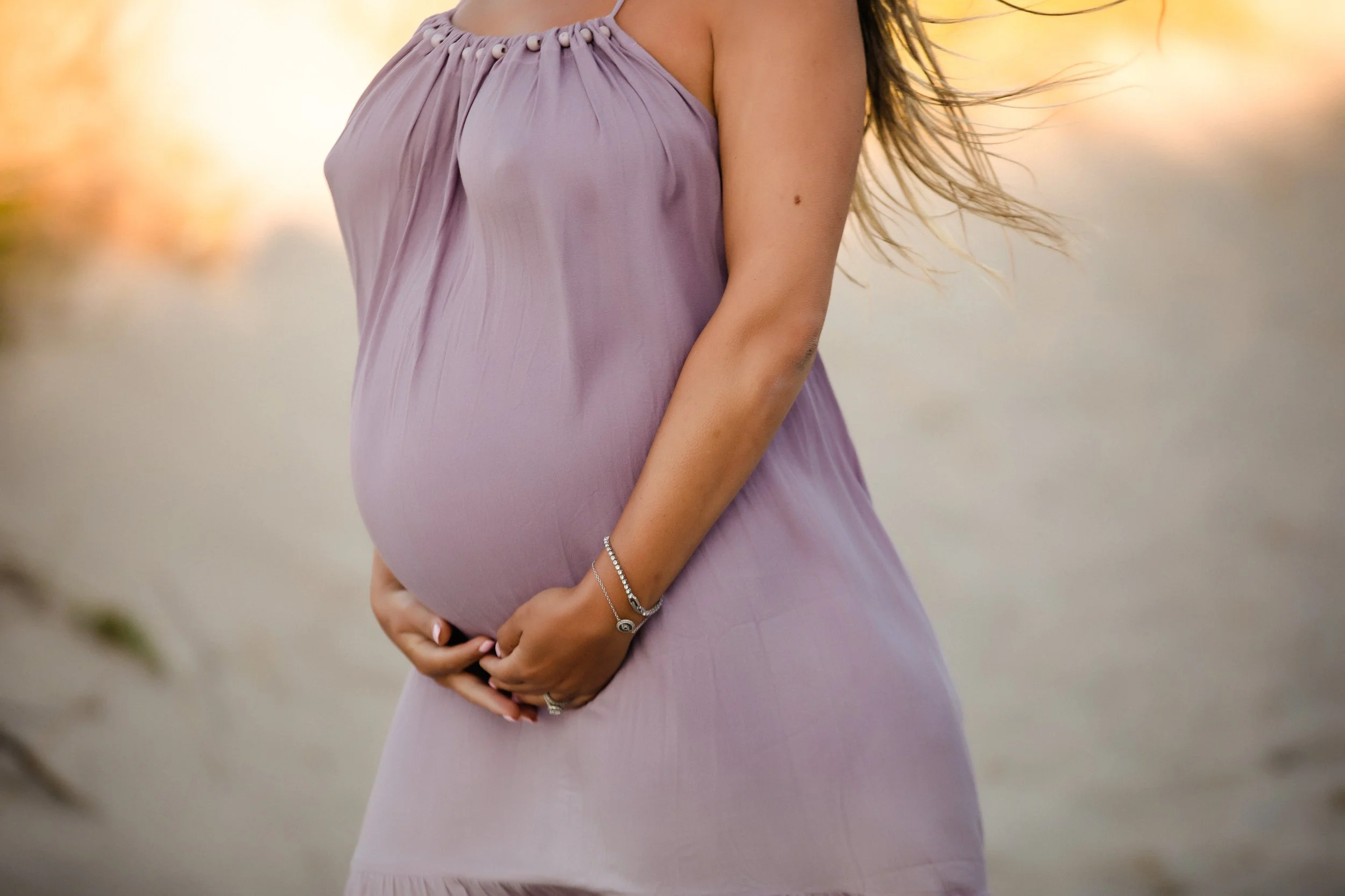 A pregnant woman in a pink dress holding her belly outdoors during sunset