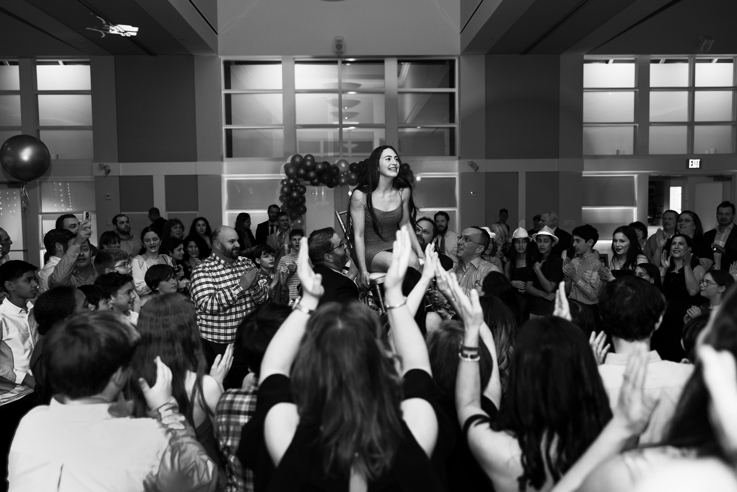A woman sitting on a chair being lifted by someone in a crowd at an indoor celebration with many people surrounding her, clapping and taking pictures.