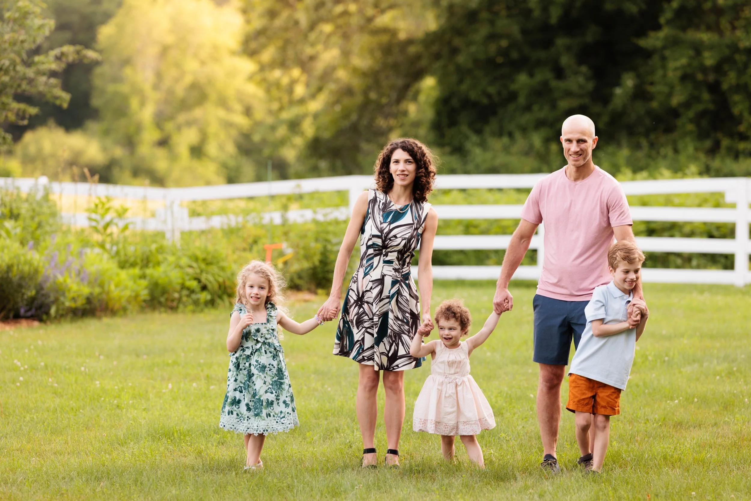 A family of two adults and three children walking together outside on a grassy area, holding hands and smiling. The background features a white fence and green trees.