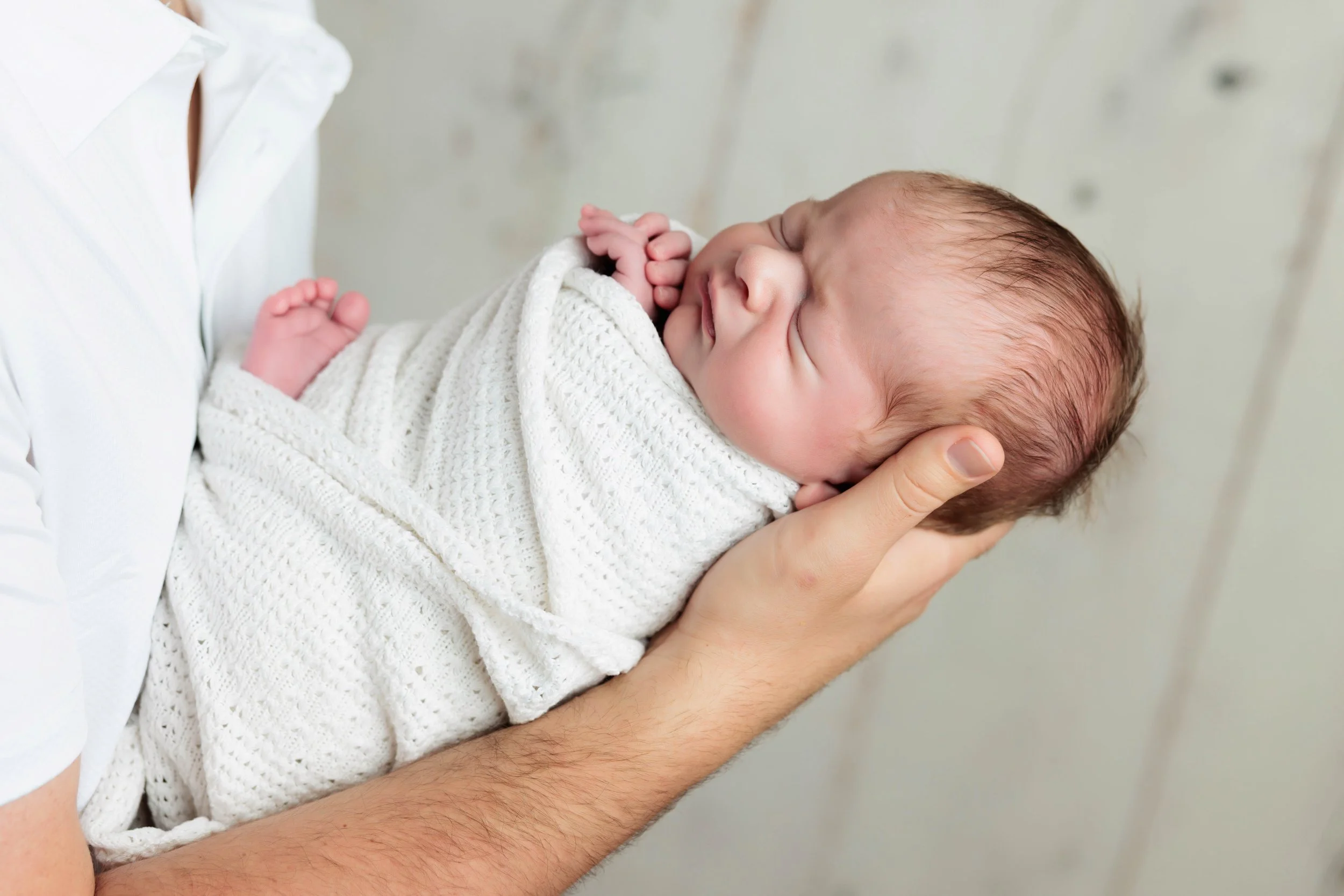 A person is holding a sleeping newborn baby wrapped in a white knit blanket, showing a peaceful expression with closed eyes, against a light-colored background.
