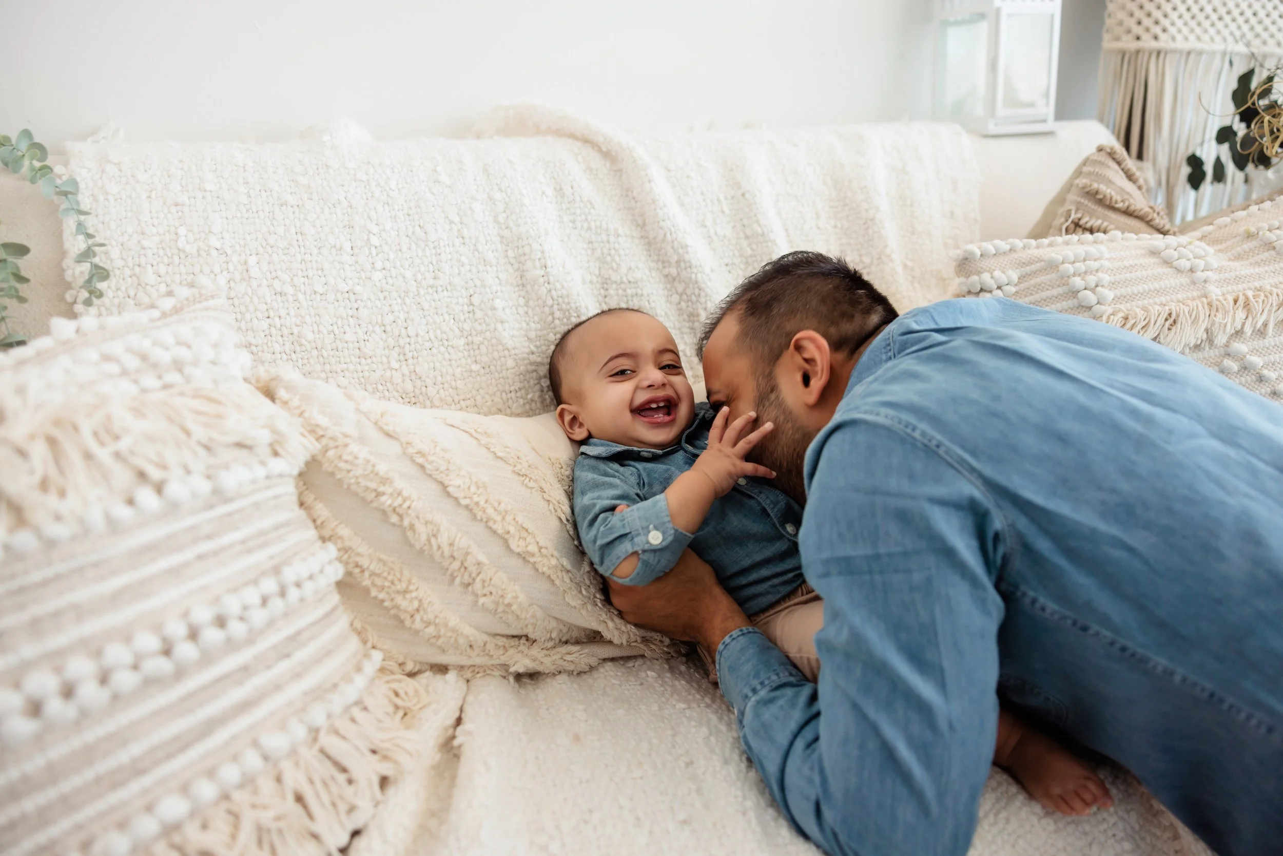 A man is playfully leaning close to a laughing baby on a cream-colored sofa in a bright living room.