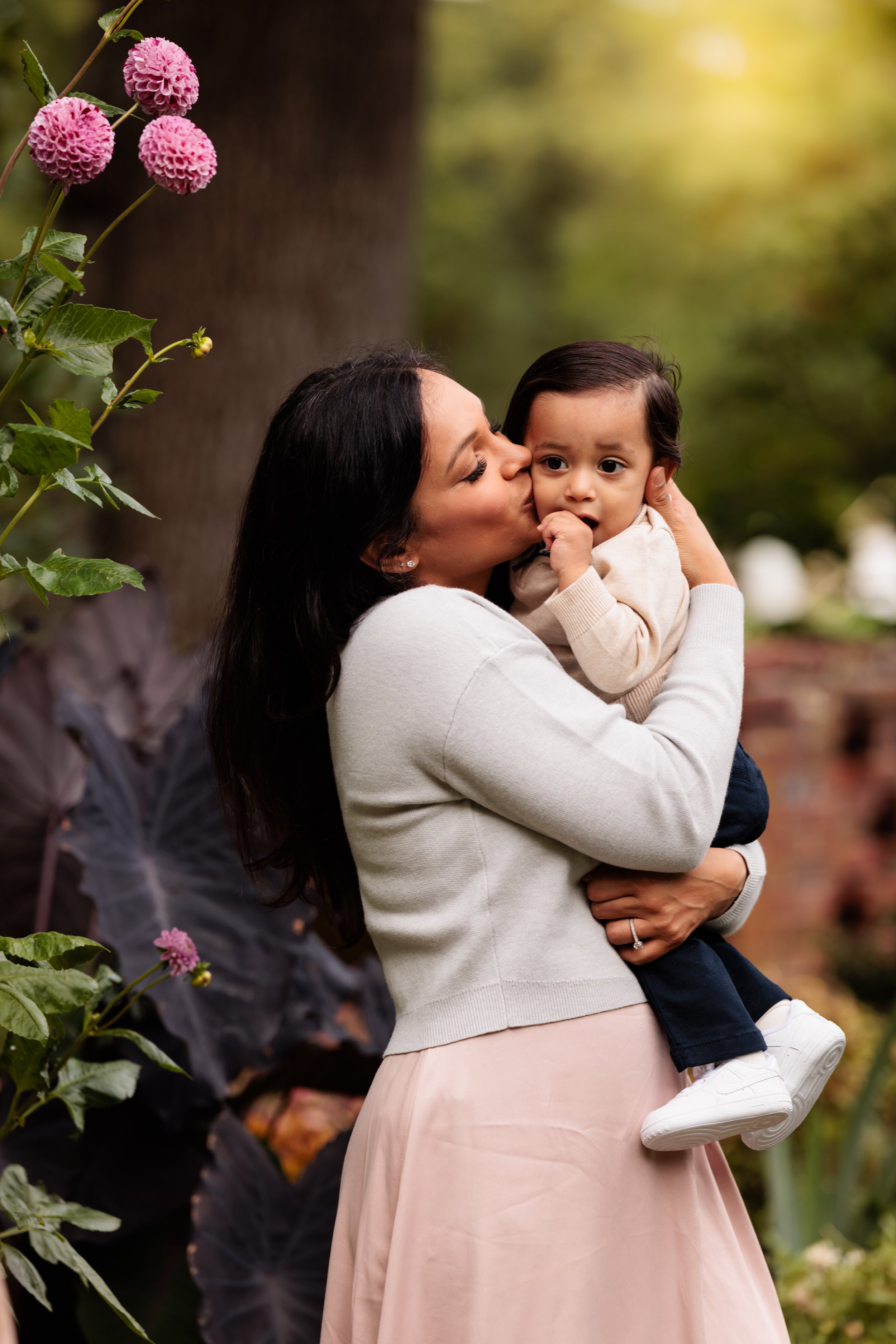 A woman holding a young boy and giving him a kiss on the cheek outdoors with greenery and pink flowers nearby.