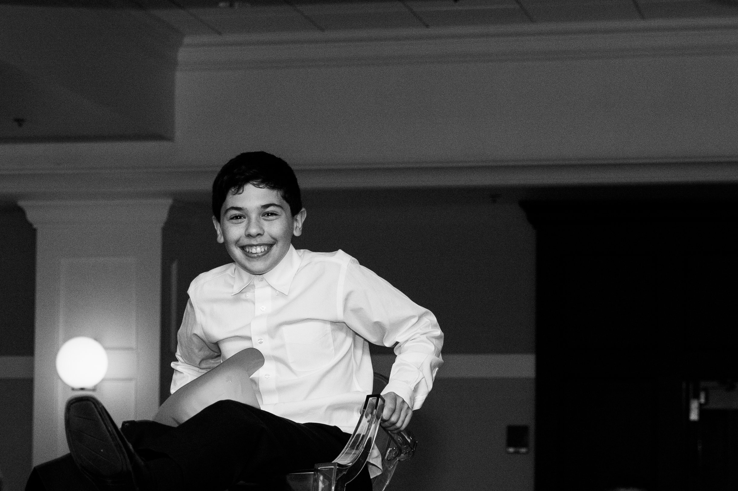 A young boy with a big smile, wearing a white button-up shirt, sitting on a chair in a room with a lit lamp in the background.