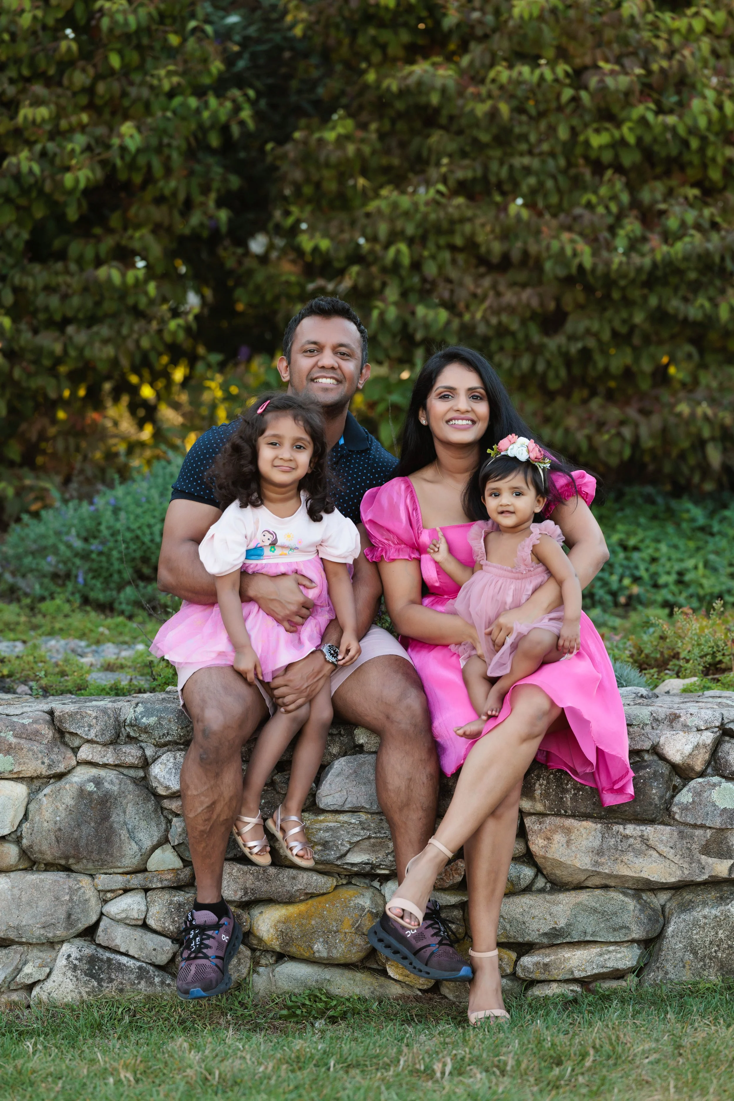 Family of four, with a man, woman, and two young girls, sitting on a stone wall outdoors. The woman and girls are dressed in pink dresses, and the man is wearing a navy blue shirt and shorts. The background features green foliage.