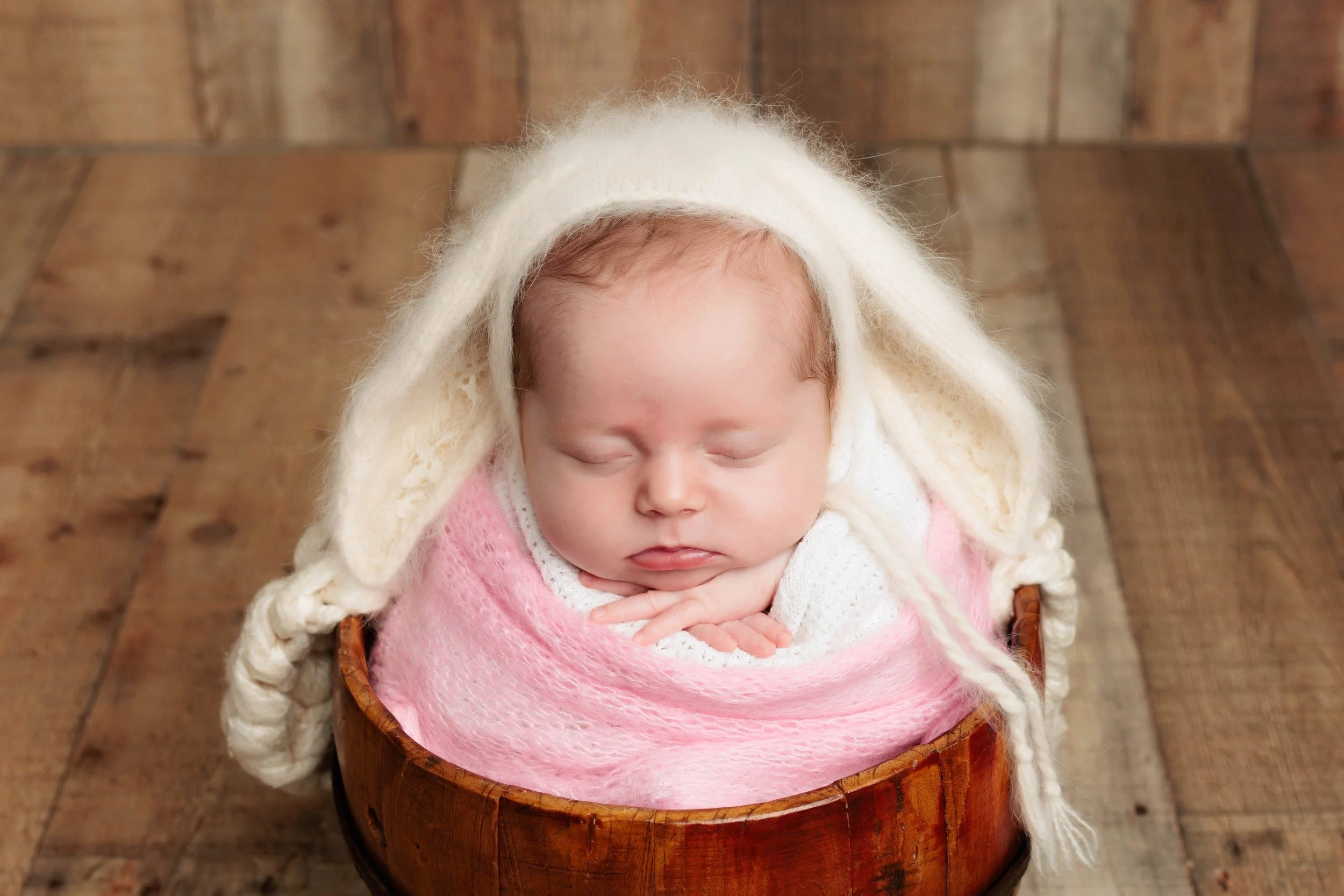 Adorable sleeping newborn baby with light skin, wrapped in a pink blanket, wearing a cream-colored sweater and with a cream-colored knitted hood draped over their head, lying in a wooden bowl on a wooden floor.