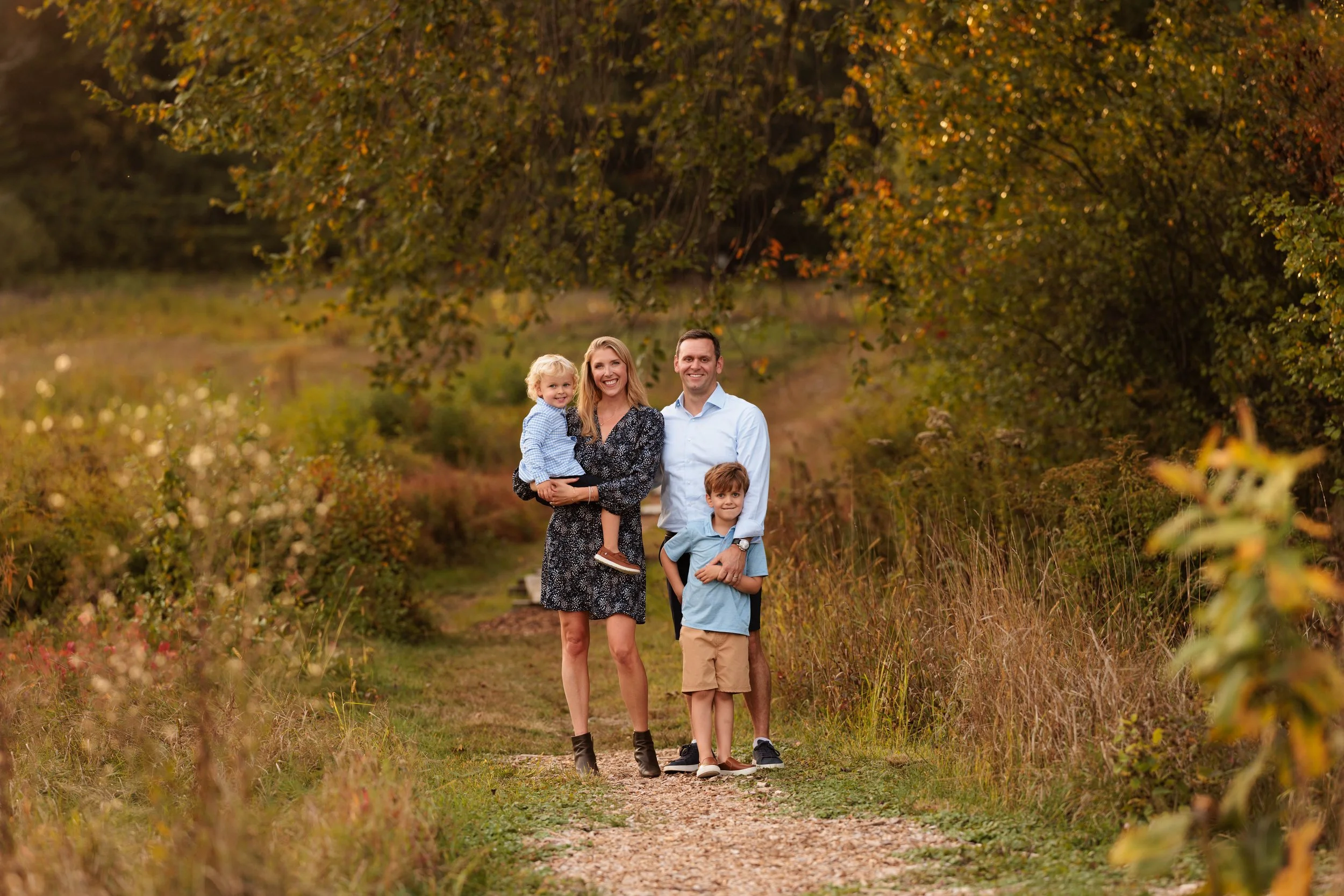 A family of four standing on a nature trail surrounded by autumn trees and foliage, smiling at the camera.
