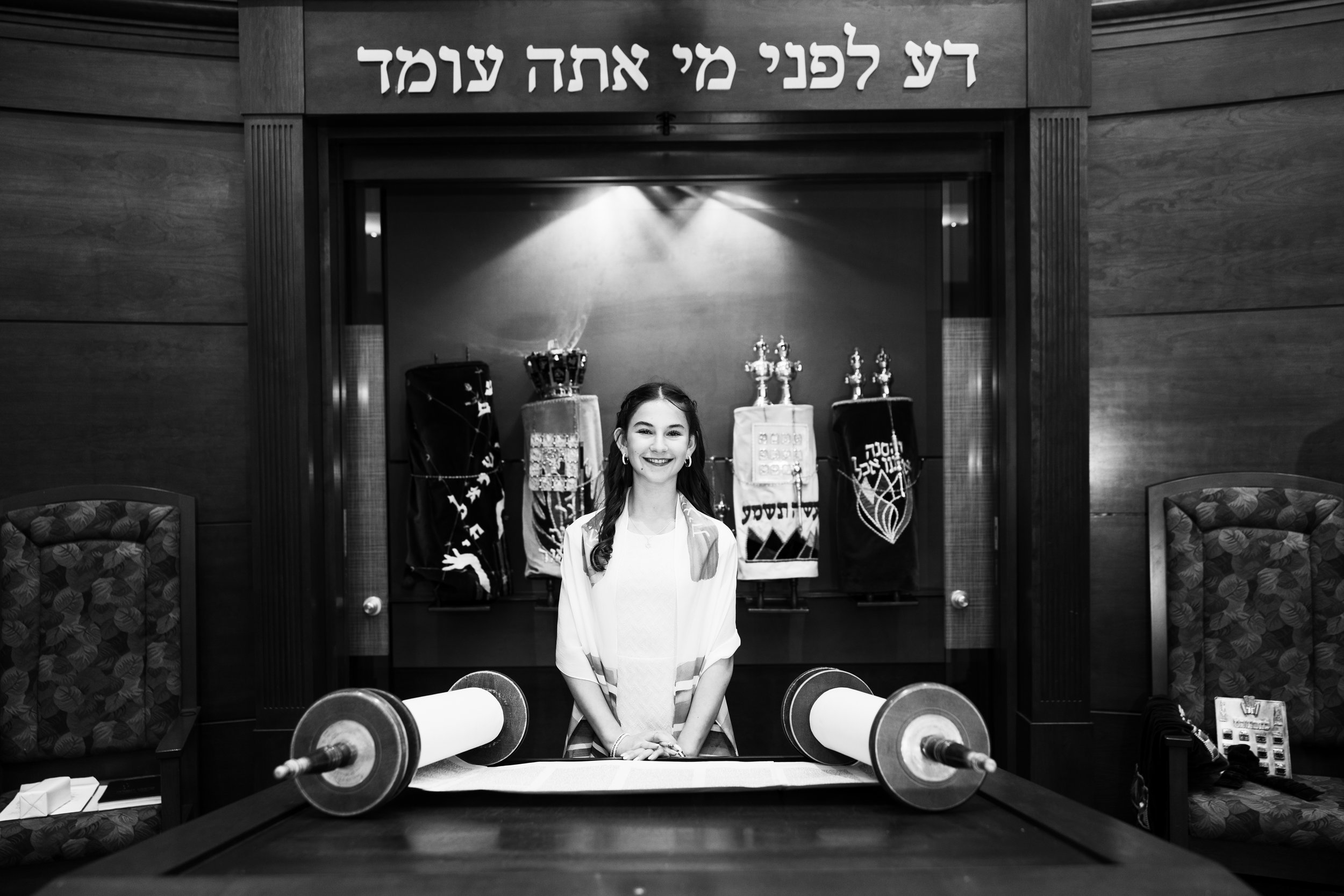 A young girl smiling behind a table with a Torah scroll, in a Jewish religious setting, with Hebrew writings above her and religious artifacts in the background.