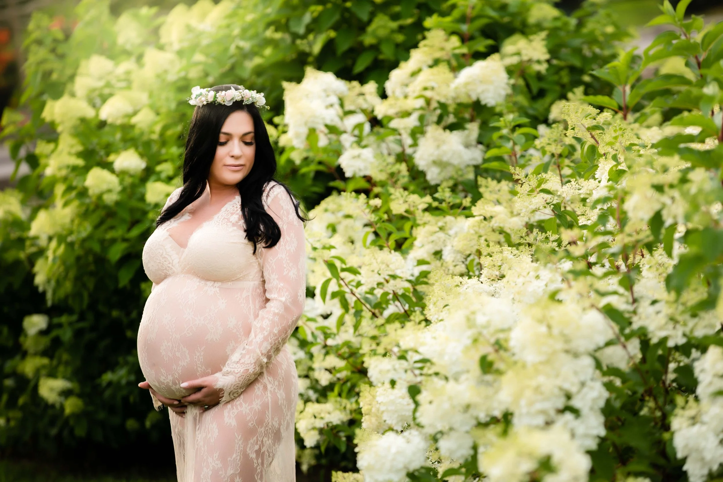Pregnant woman in a lace dress standing amidst white flowers with a floral crown on her head, cradling her belly.
