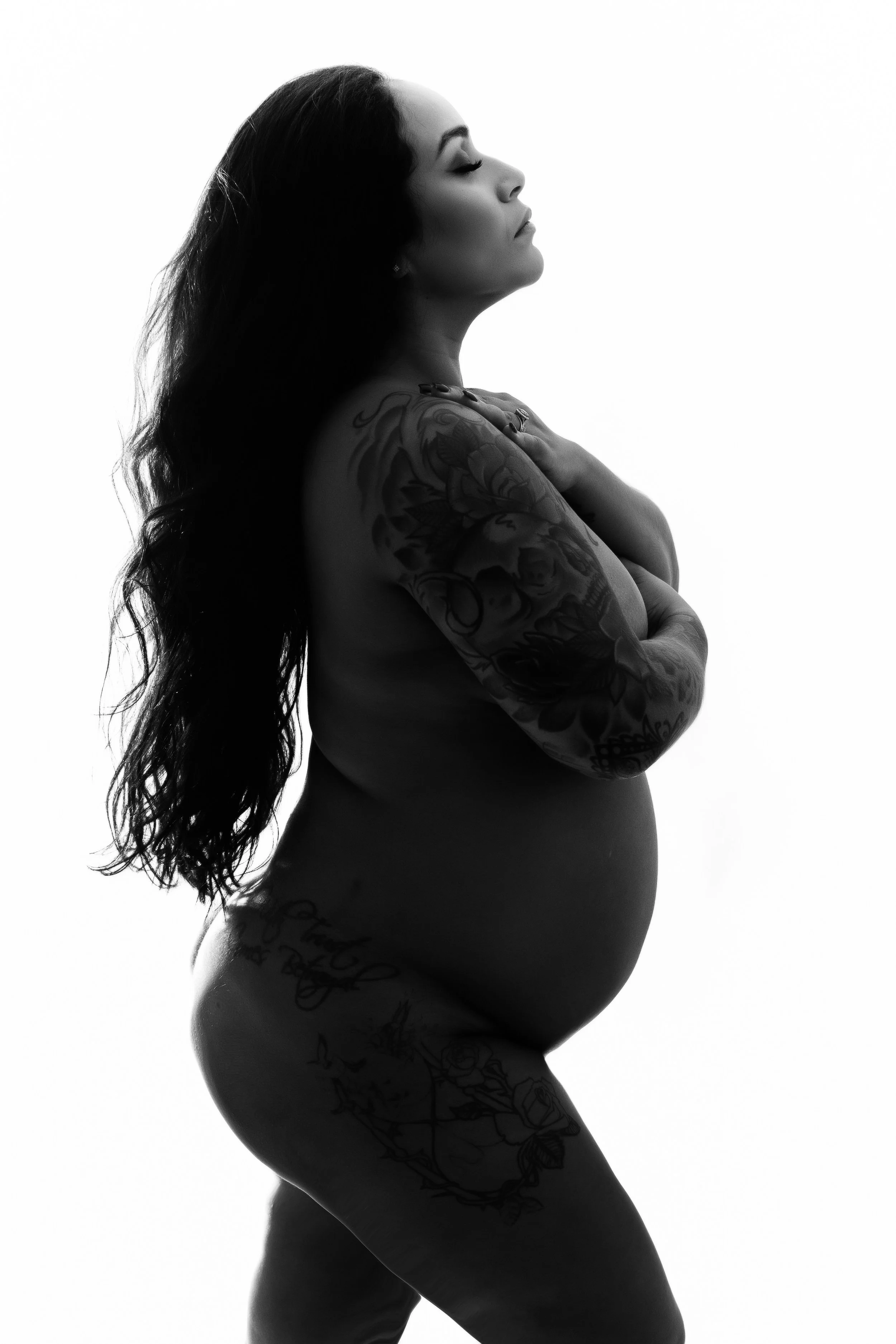 Black and white side profile of a pregnant woman with long, wavy hair, closed eyes, tattoos on her arms, shoulder, and hip, standing with her hands crossed over her chest.