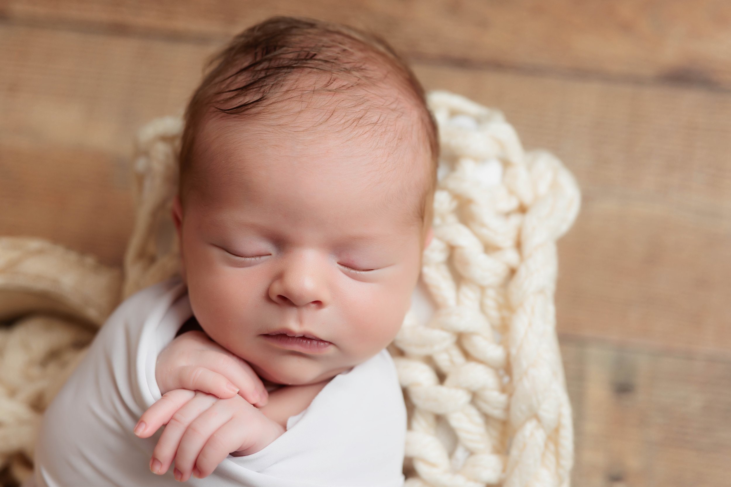 Close-up of a sleeping baby with hands folded near the face, lying on a soft, cream-colored blanket on a wooden surface.