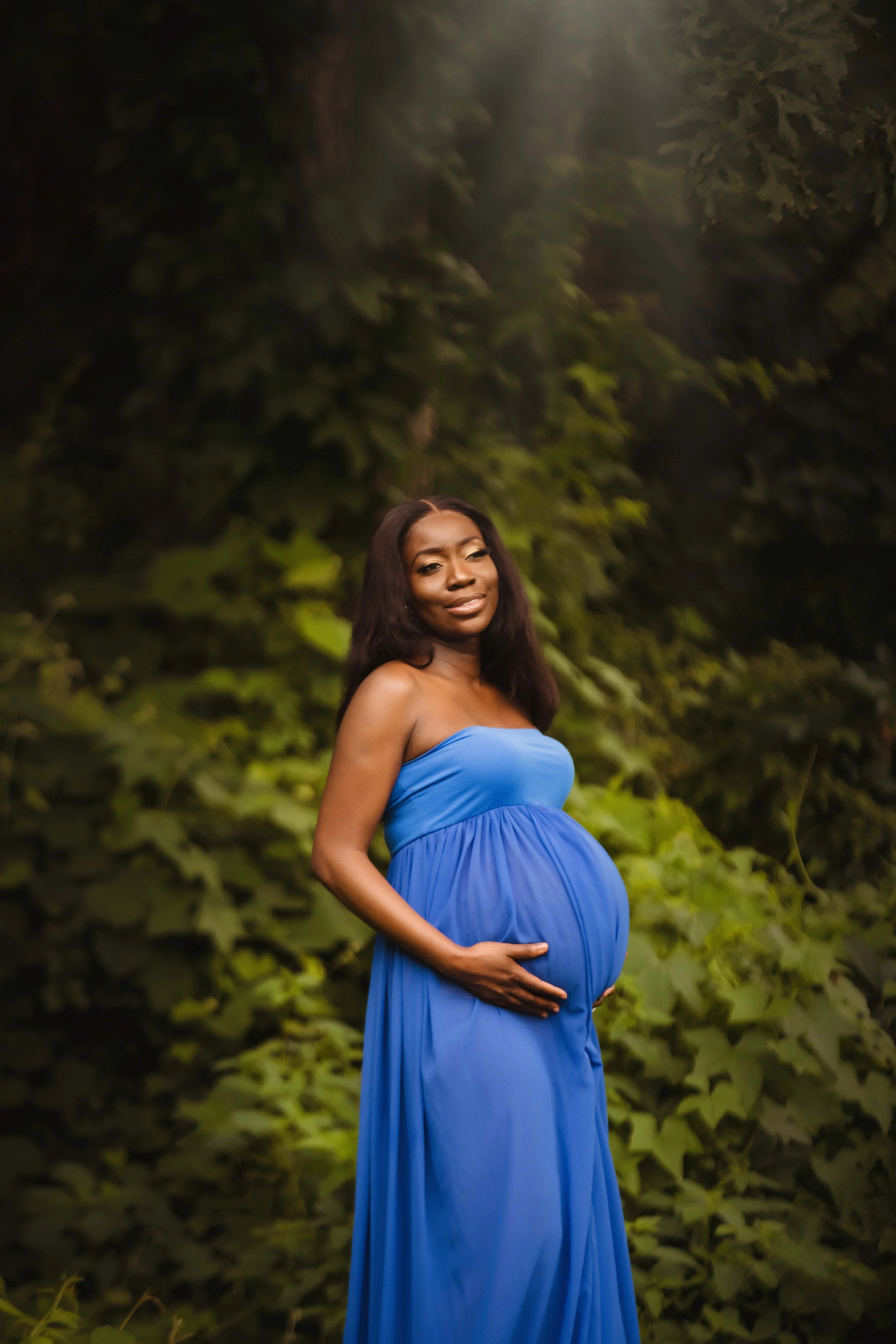 A pregnant woman wearing a blue strapless dress standing outdoors among green foliage.