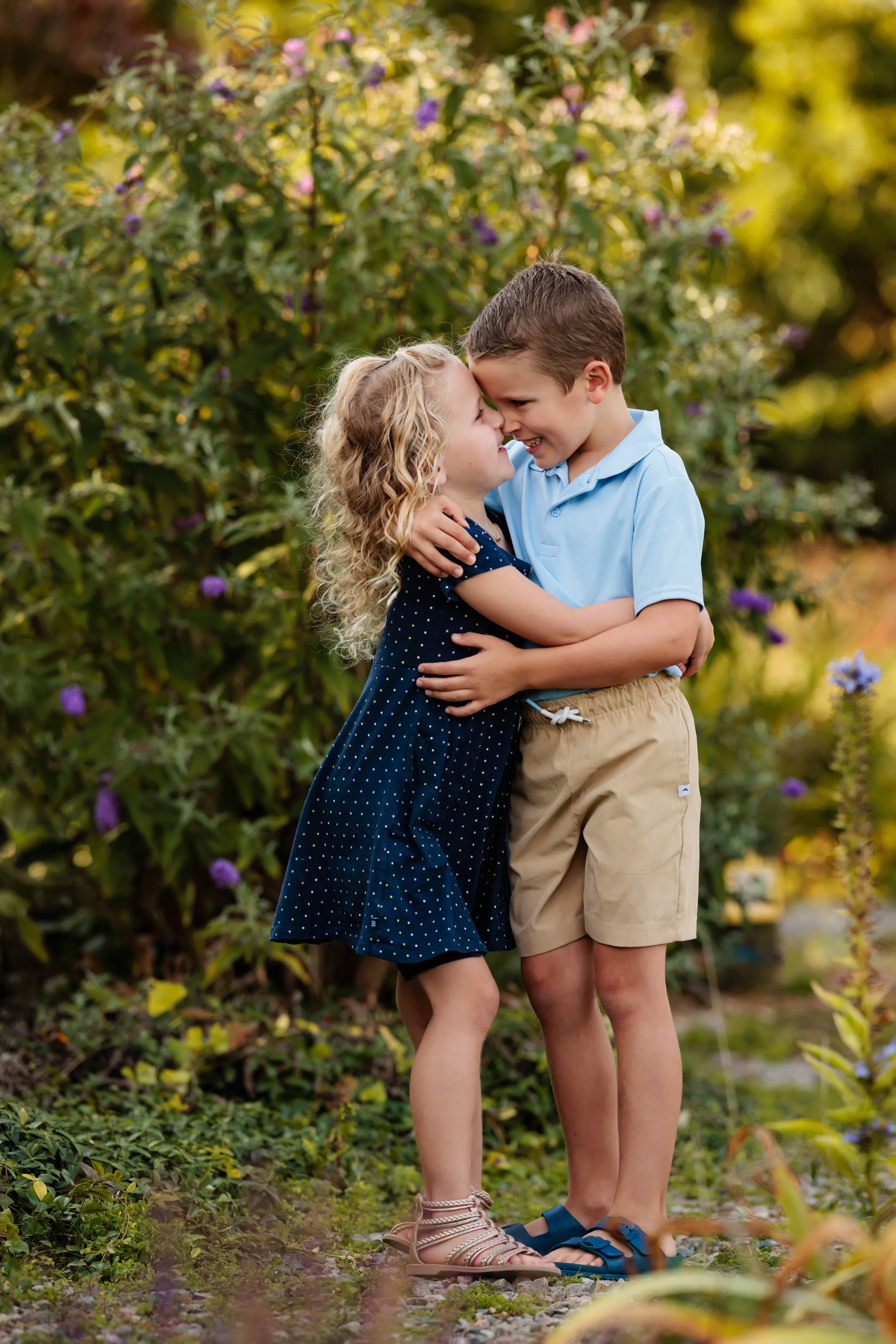 A young boy and girl joyfully embrace outdoors in front of a bush with purple flowers, smiling and touching noses.
