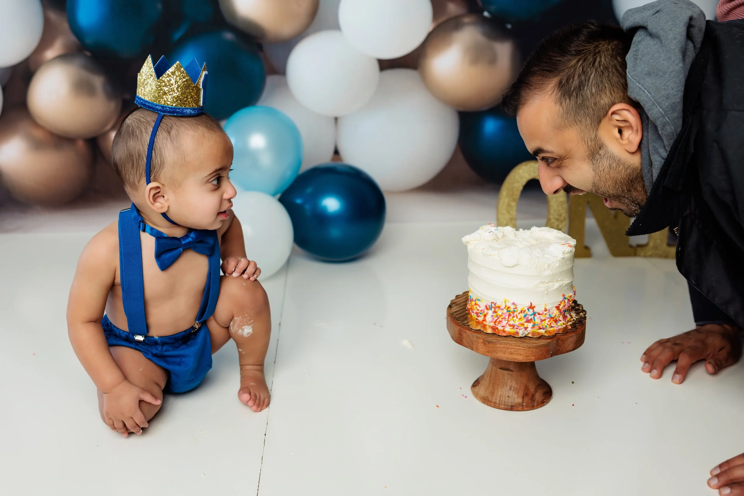 A baby boy wearing a blue suspenders and a bow tie, with a birthday crown on his head, sitting on the floor. He is looking at a man who is leaning towards a birthday cake with white frosting and colorful sprinkles. The background has balloons in whit