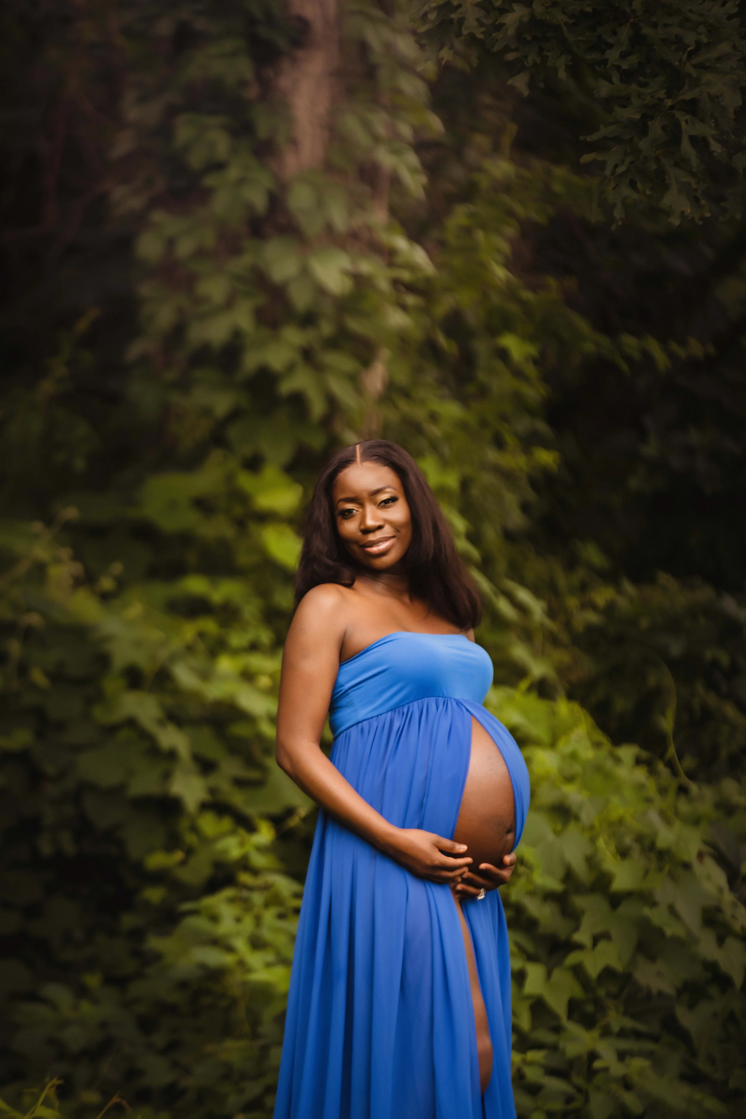 A pregnant woman in a strapless blue dress standing outdoors in front of dense green foliage, gently holding her belly and smiling at the camera.