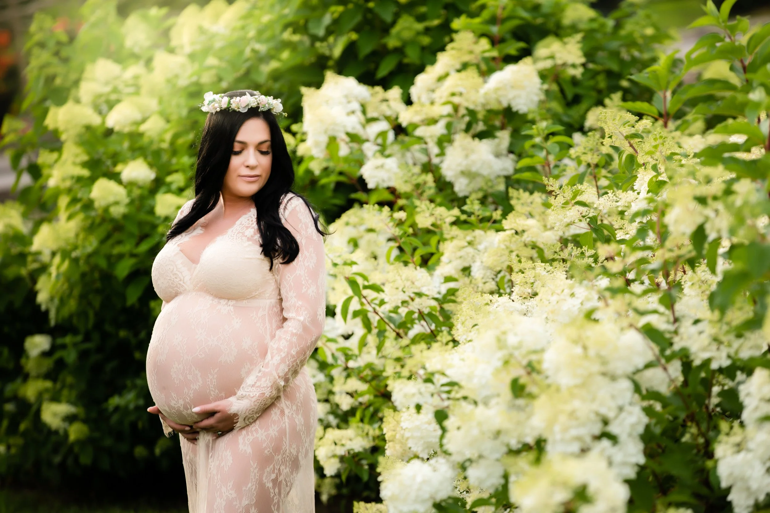 Pregnant woman with long dark hair wearing a light pink lace dress, standing in a garden with white hydrangea flowers, looking down with her hands on her belly.
