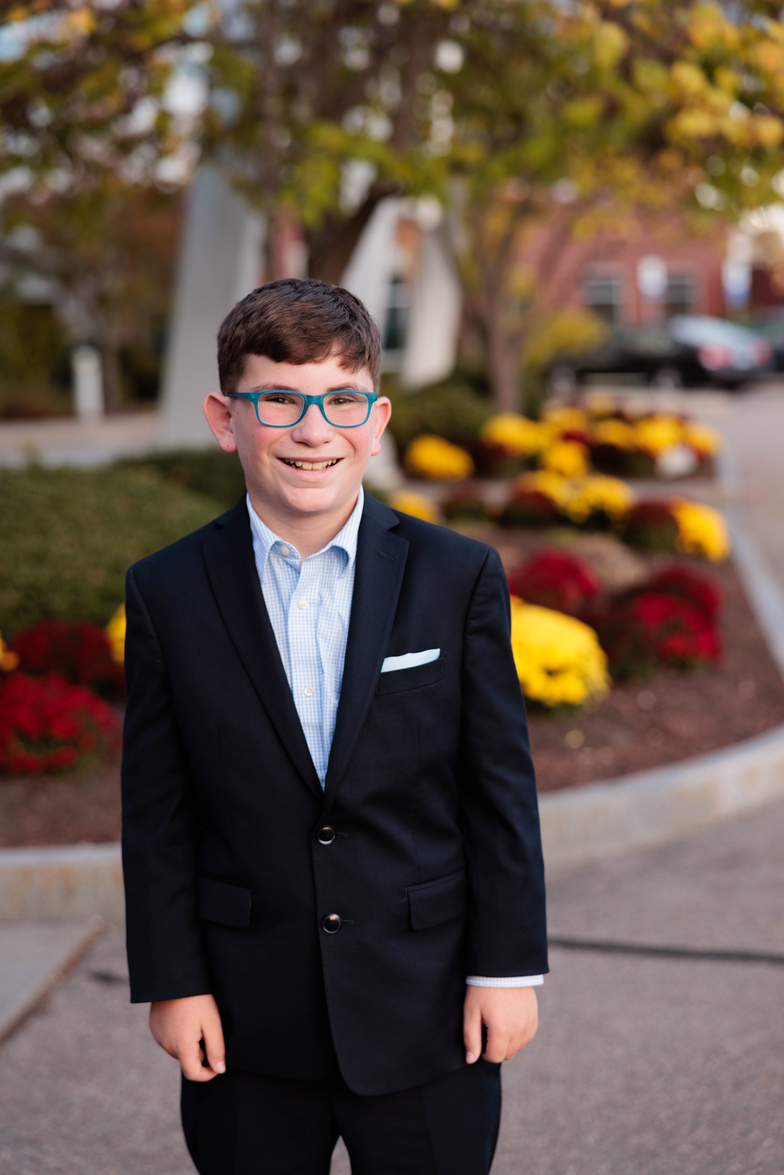 A young boy with glasses and a big smile, dressed in a black blazer and light blue shirt, standing outdoors with a background of colorful fall flowers and trees.