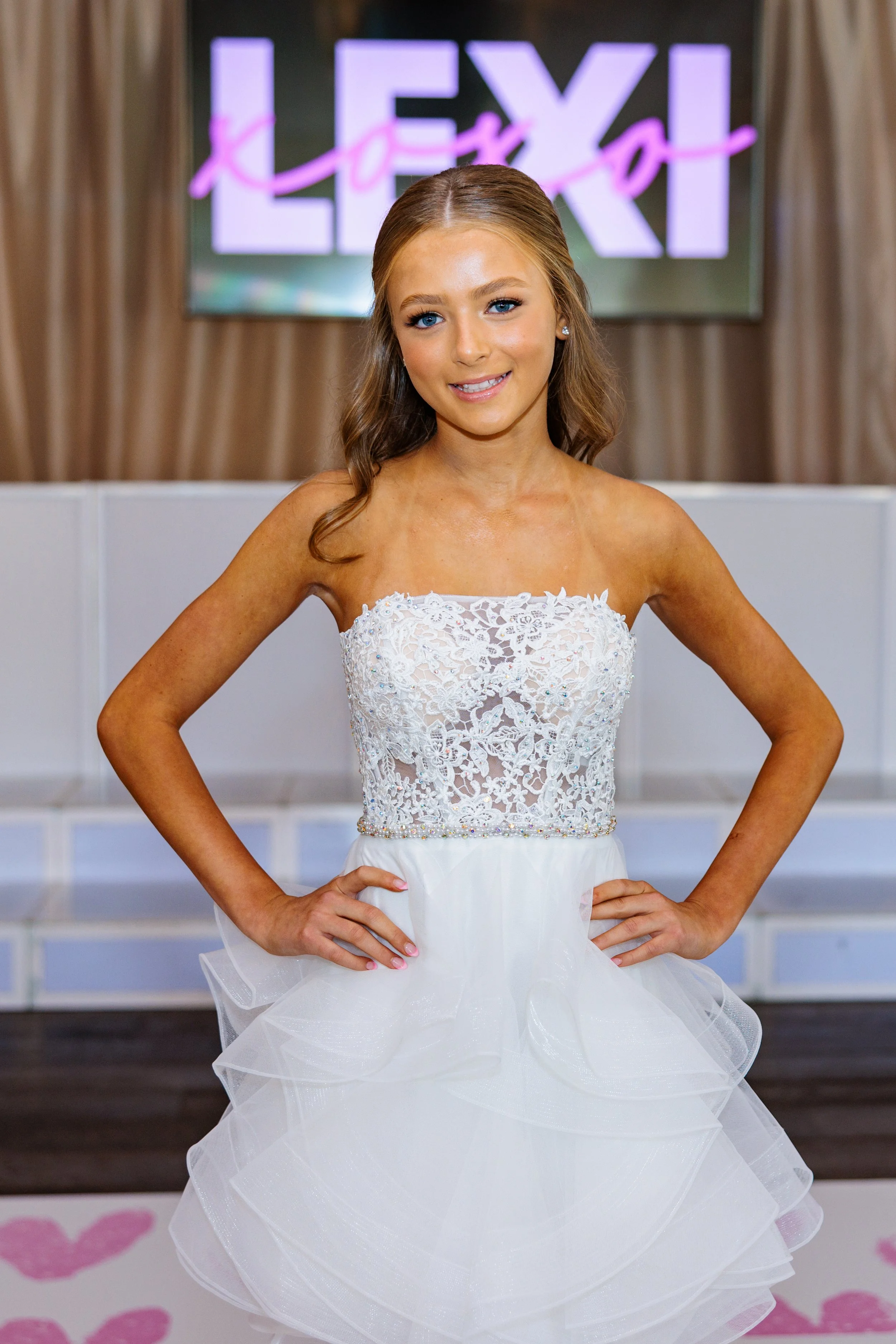 Young woman in a white lace strapless dress with a full tulle skirt, posing with hands on hips in front of a blurred background with the word "LEXY" on a screen.
