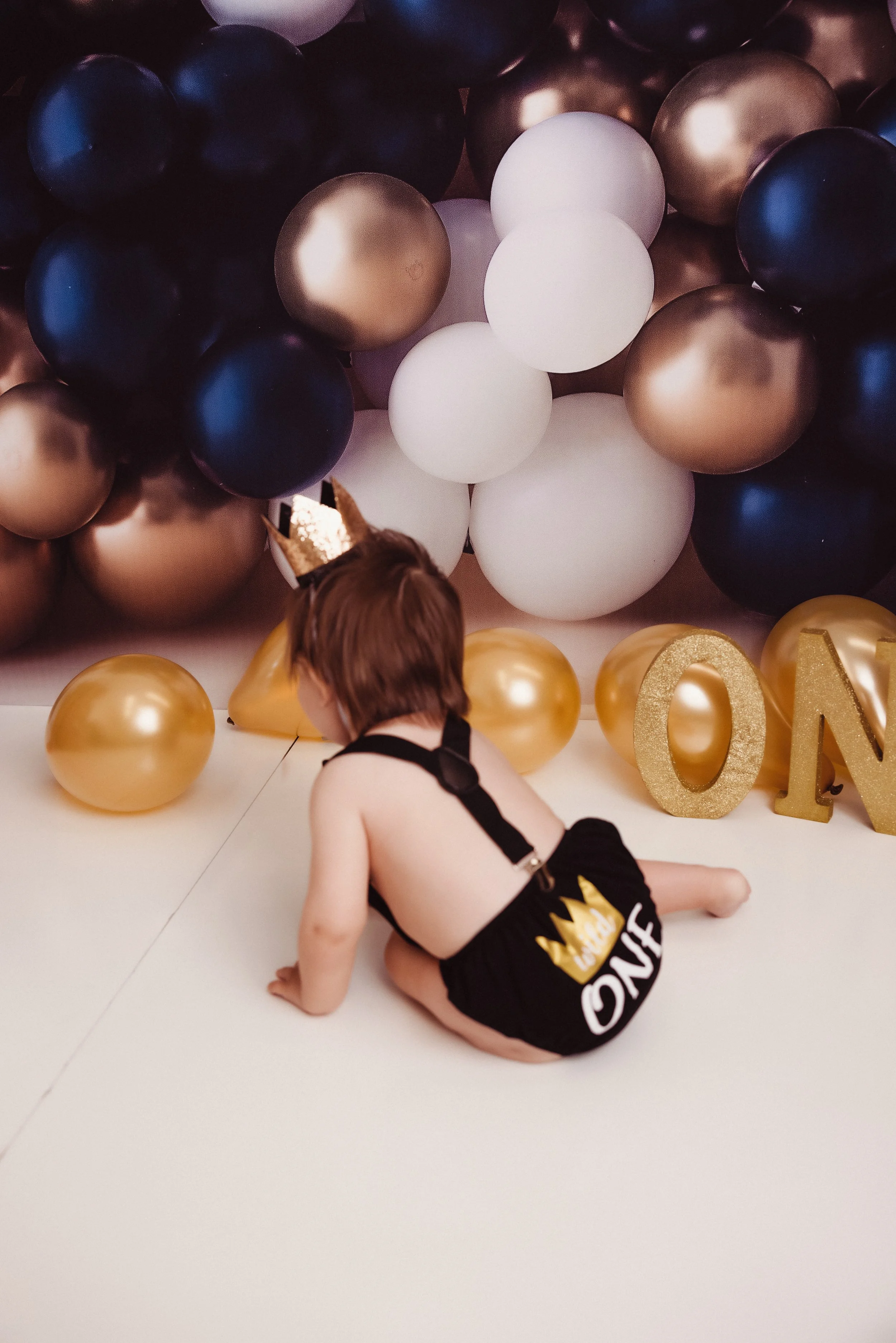 A young child with a golden crown headband, wearing a black diaper with a gold crown and the word 'KING' on the back, sitting on the floor in front of a backdrop of black, white, and gold balloons, and gold 'ONE' letters.