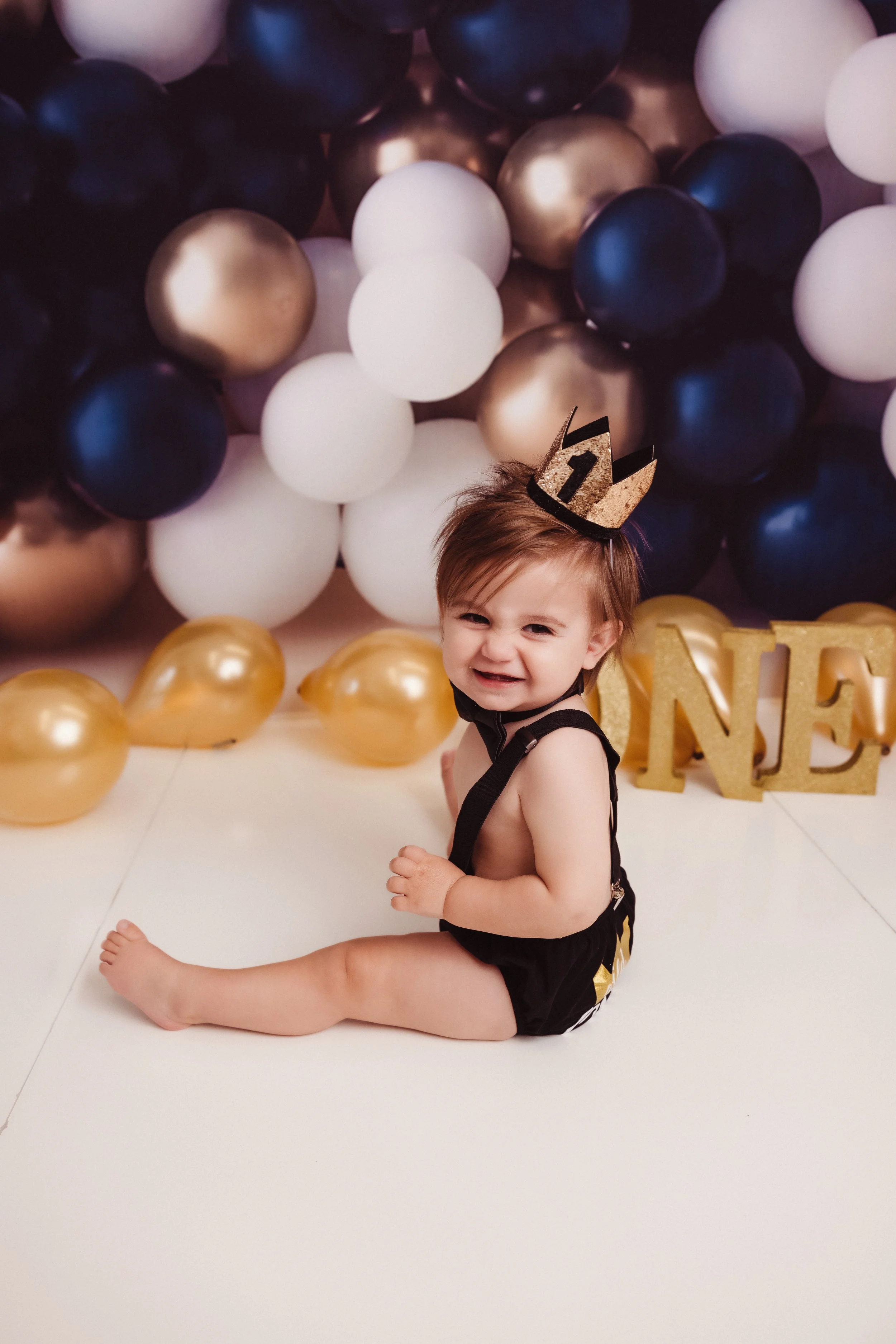 A young child sitting on a white floor with balloons and a golden 'ONE' decoration in the background, wearing a birthday crown and a black outfit, celebrating their first birthday.