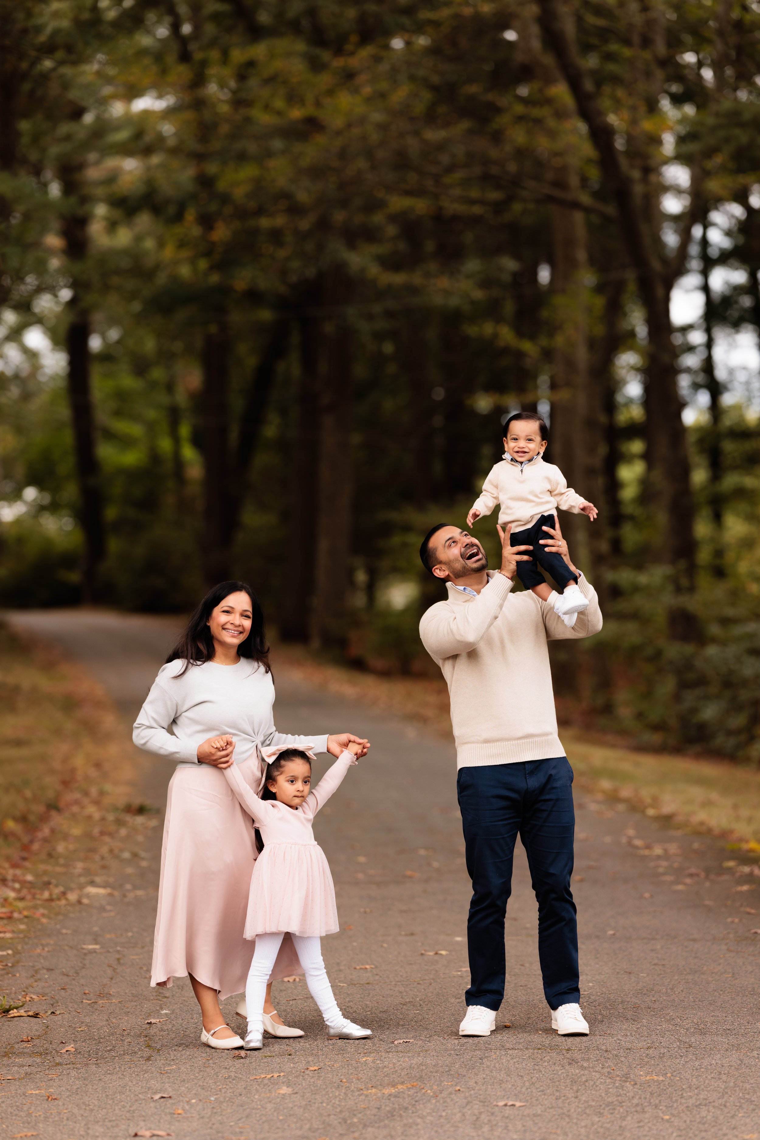 A family of four enjoying a walk in a park with autumn foliage. The father is lifting a young boy in the air, both smiling. The mother and a young girl are standing beside them, with the girl holding the mother's hand.