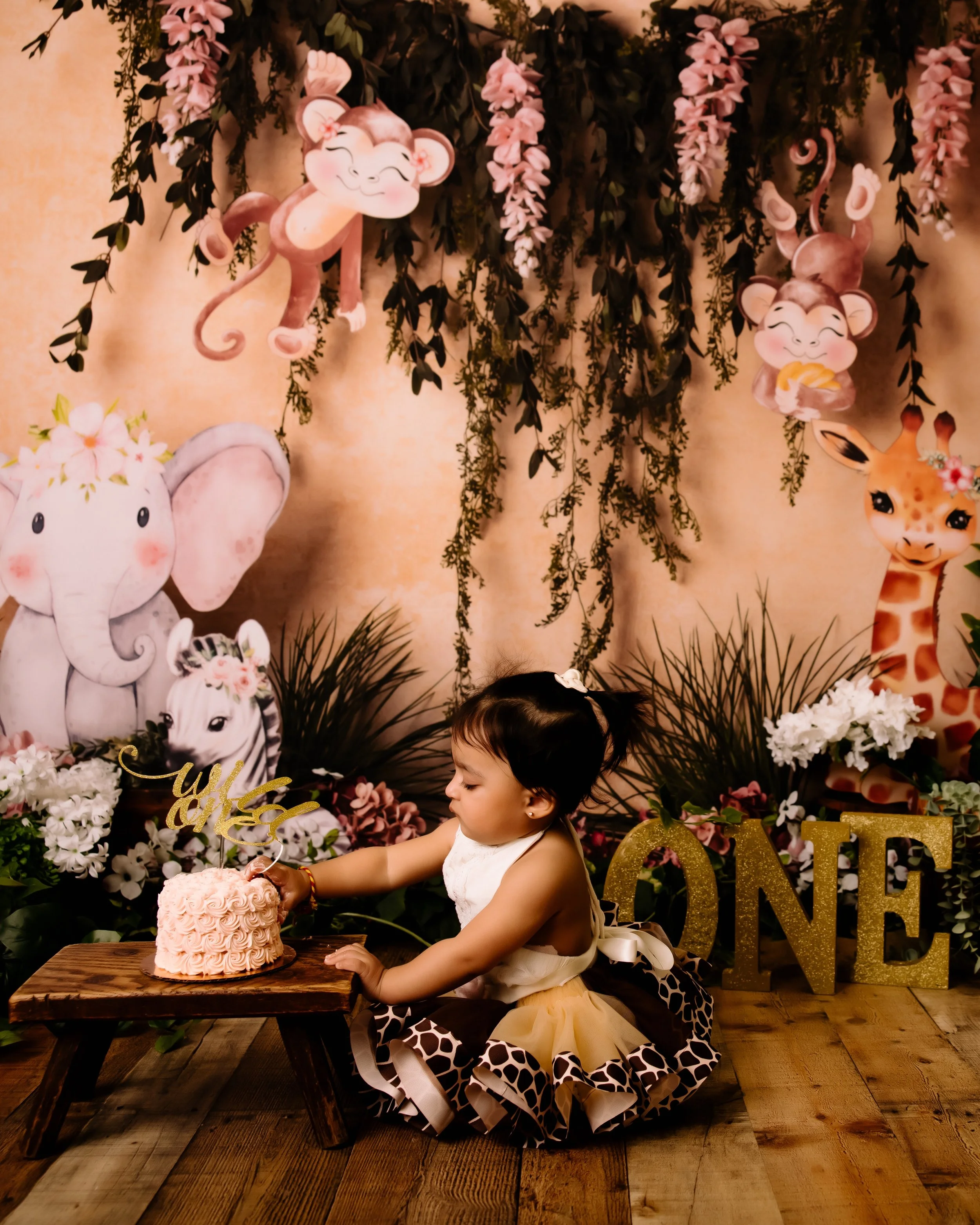 A young girl is sitting on a wooden floor at her one-year birthday celebration. She is reaching out to touch a pink cake on a small table. The background features colorful jungle animal decorations, including elephants, giraffes, and monkeys, with ha