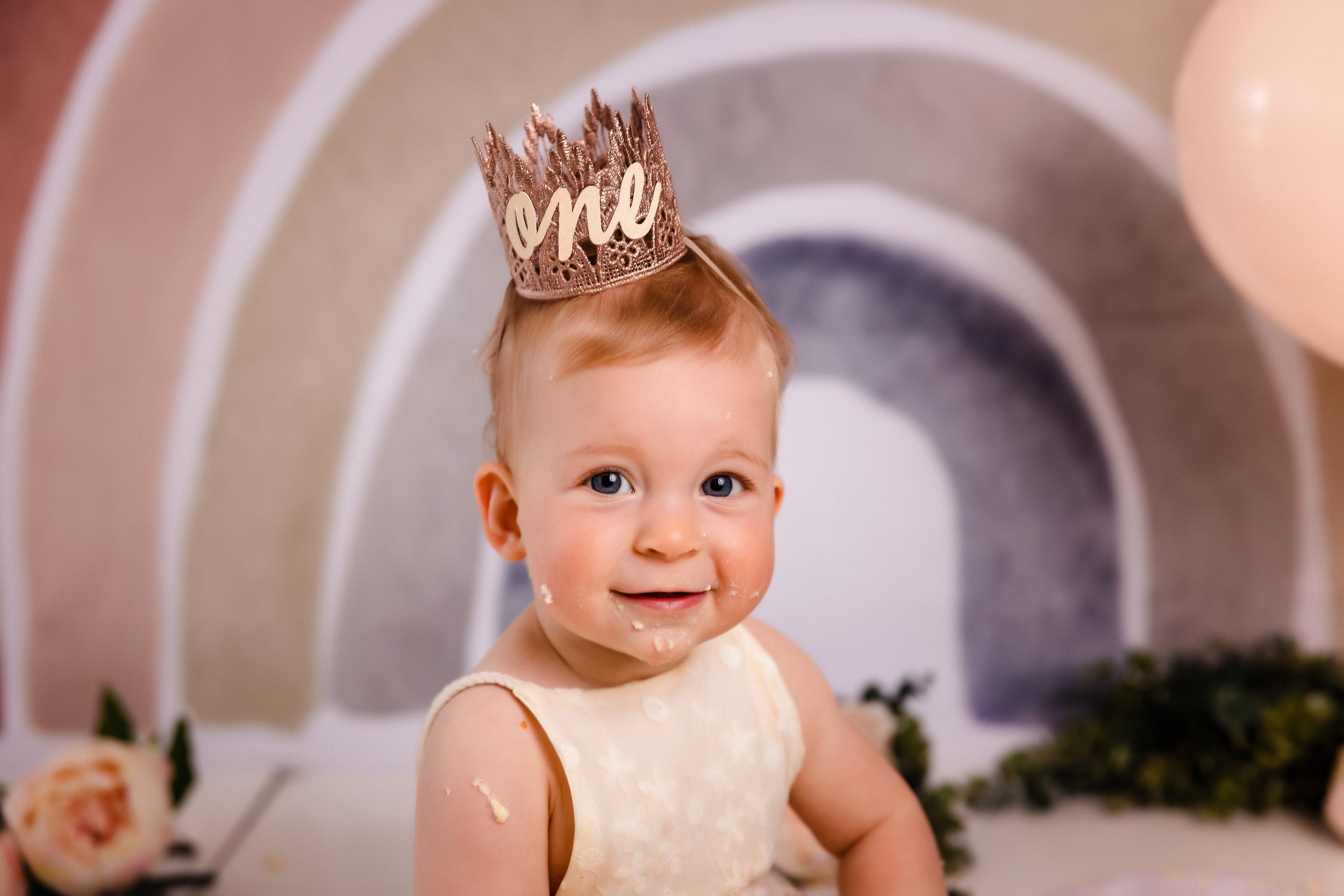 A young child celebrating a birthday, wearing a crown with "one" written on it, has cake or frosting on her face, and is smiling.