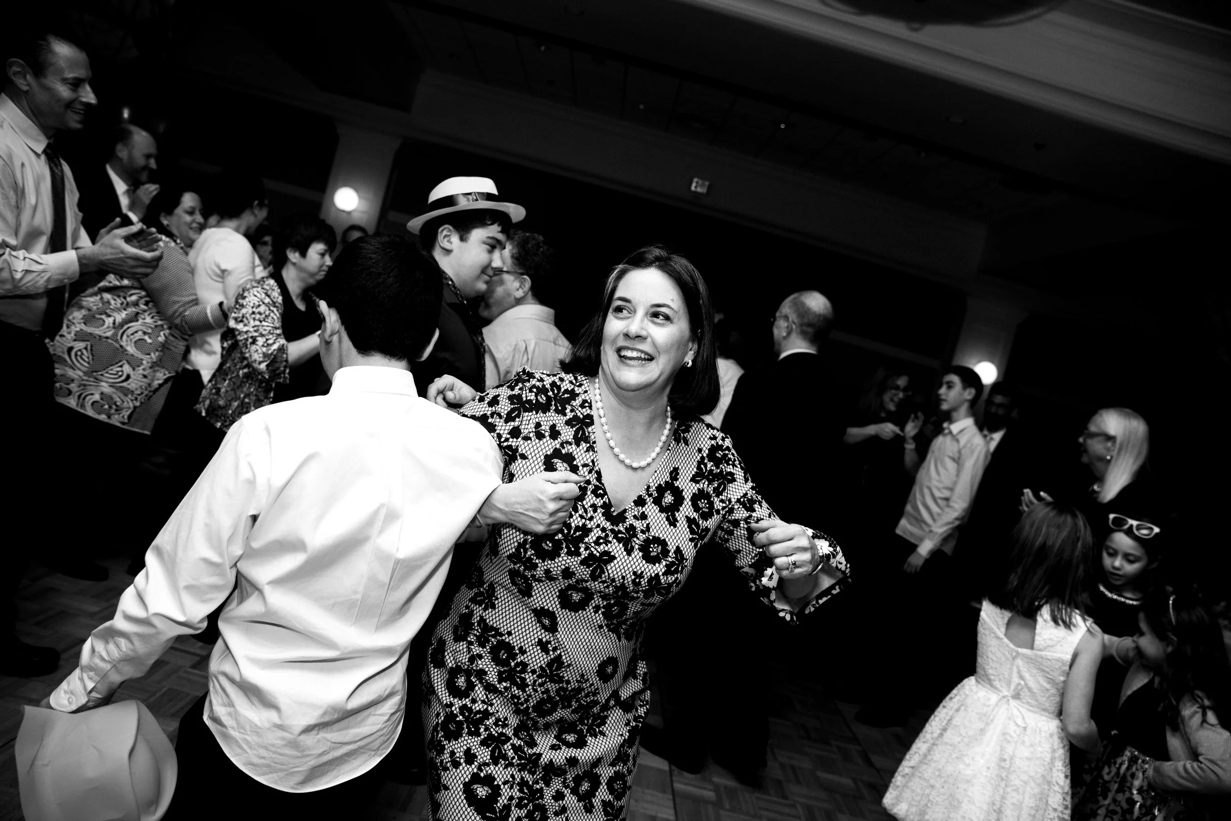 People dancing and socializing at a party or celebration in a ballroom with a wooden floor. A woman with short dark hair and a patterned dress smiles as she holds hands with a boy in a white shirt, with other guests in the background.