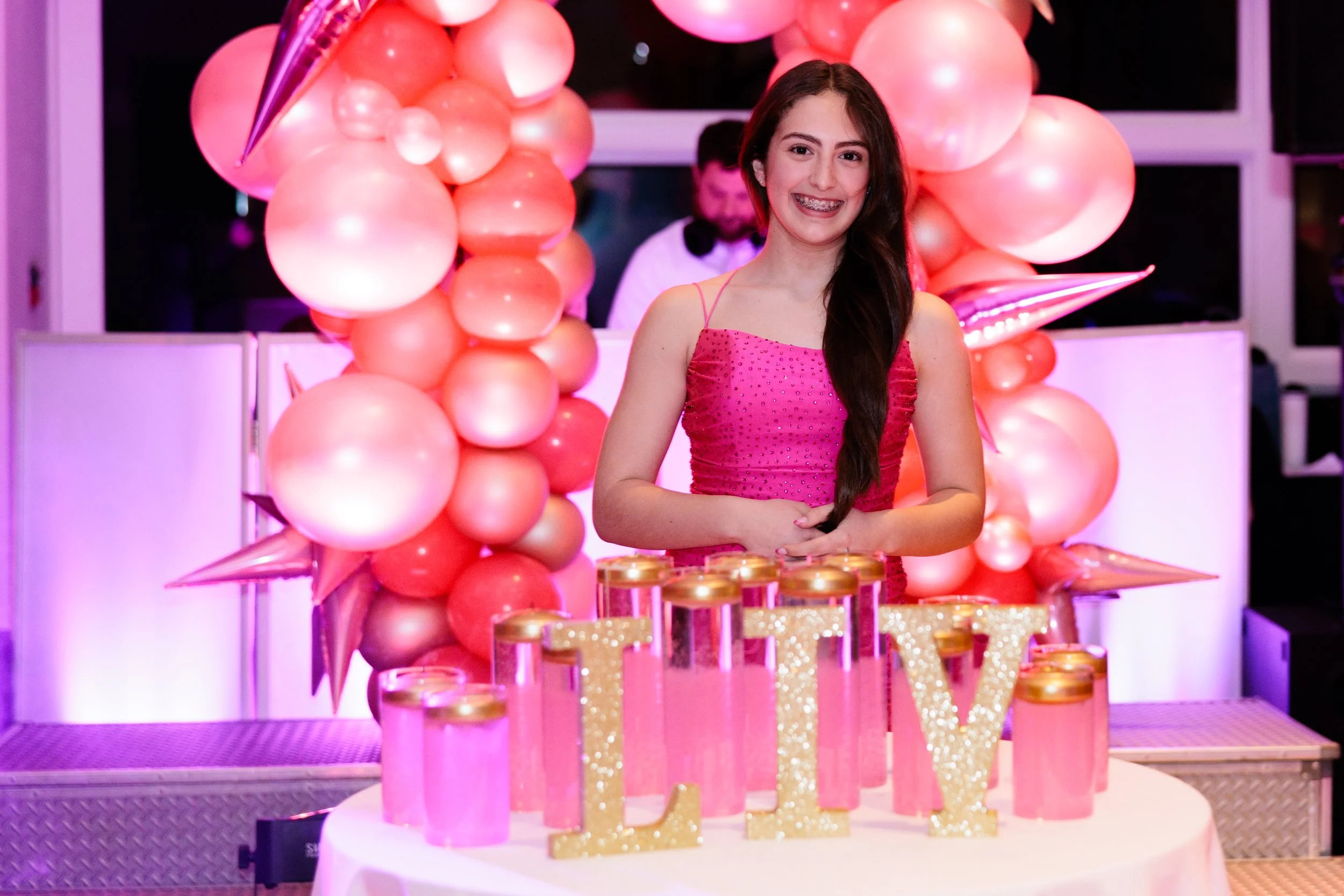 A girl smiling at a birthday party surrounded by pink balloons and decorations, with a table featuring gold-topped jars and illuminated letters spelling 'LOVE'.