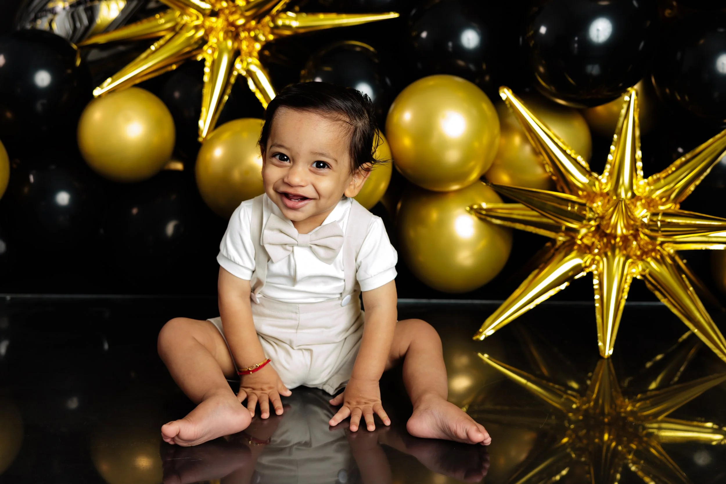 A young child sitting on a black shiny floor, smiling and looking at the camera, surrounded by gold and black balloons and large metallic gold star-shaped balloons in the background.