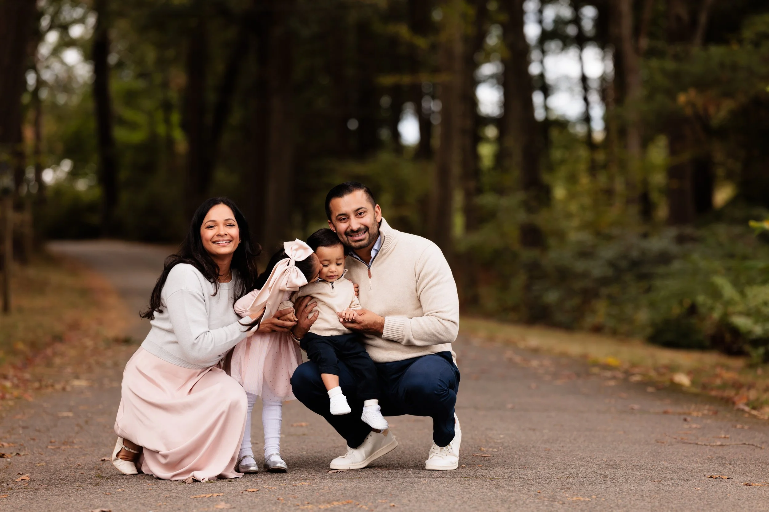 A family of four smiling outdoors on a wooded path, with a woman, a man, and two children.