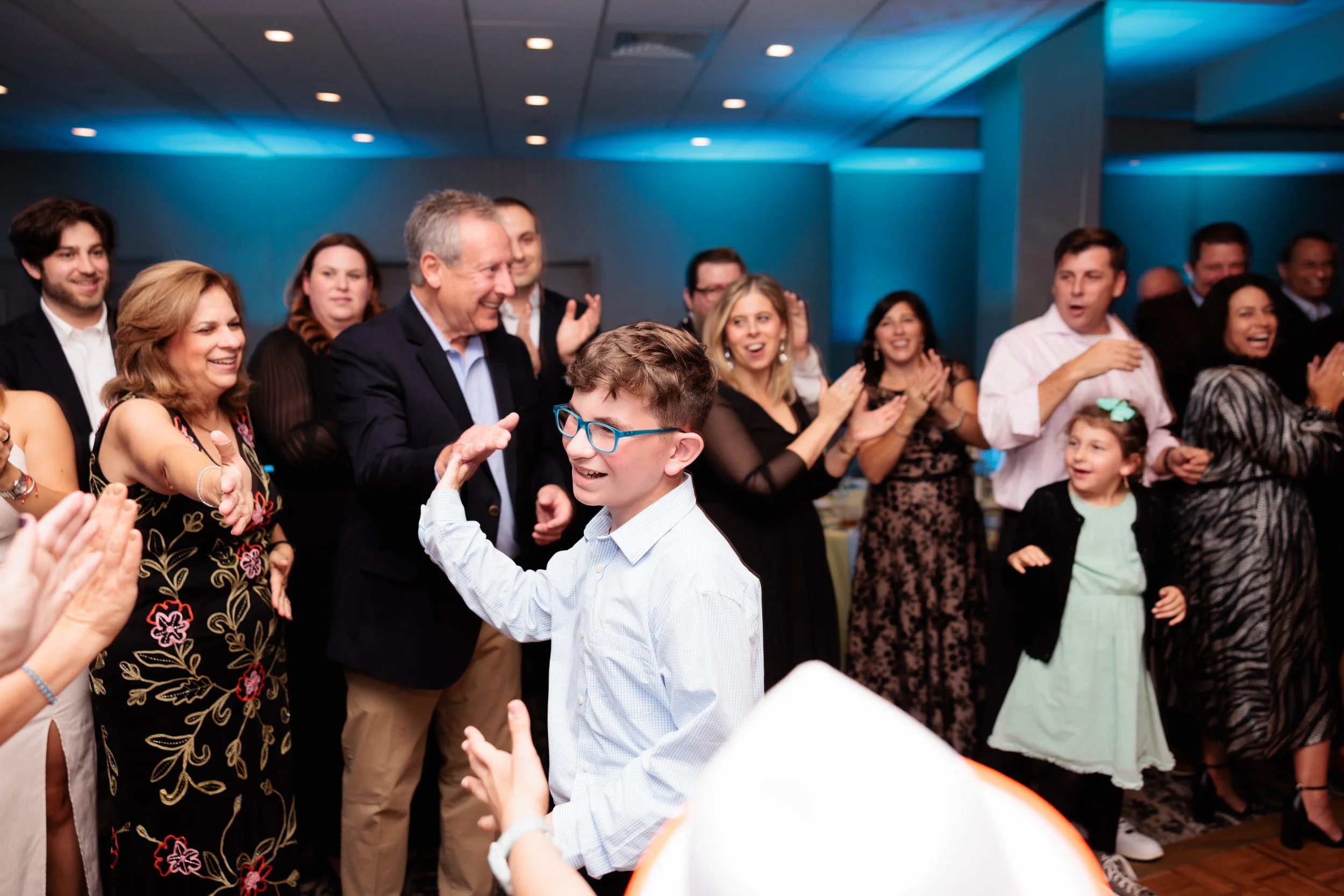 People celebrating at a party, clapping, and smiling with a boy wearing glasses and a white shirt in the foreground.