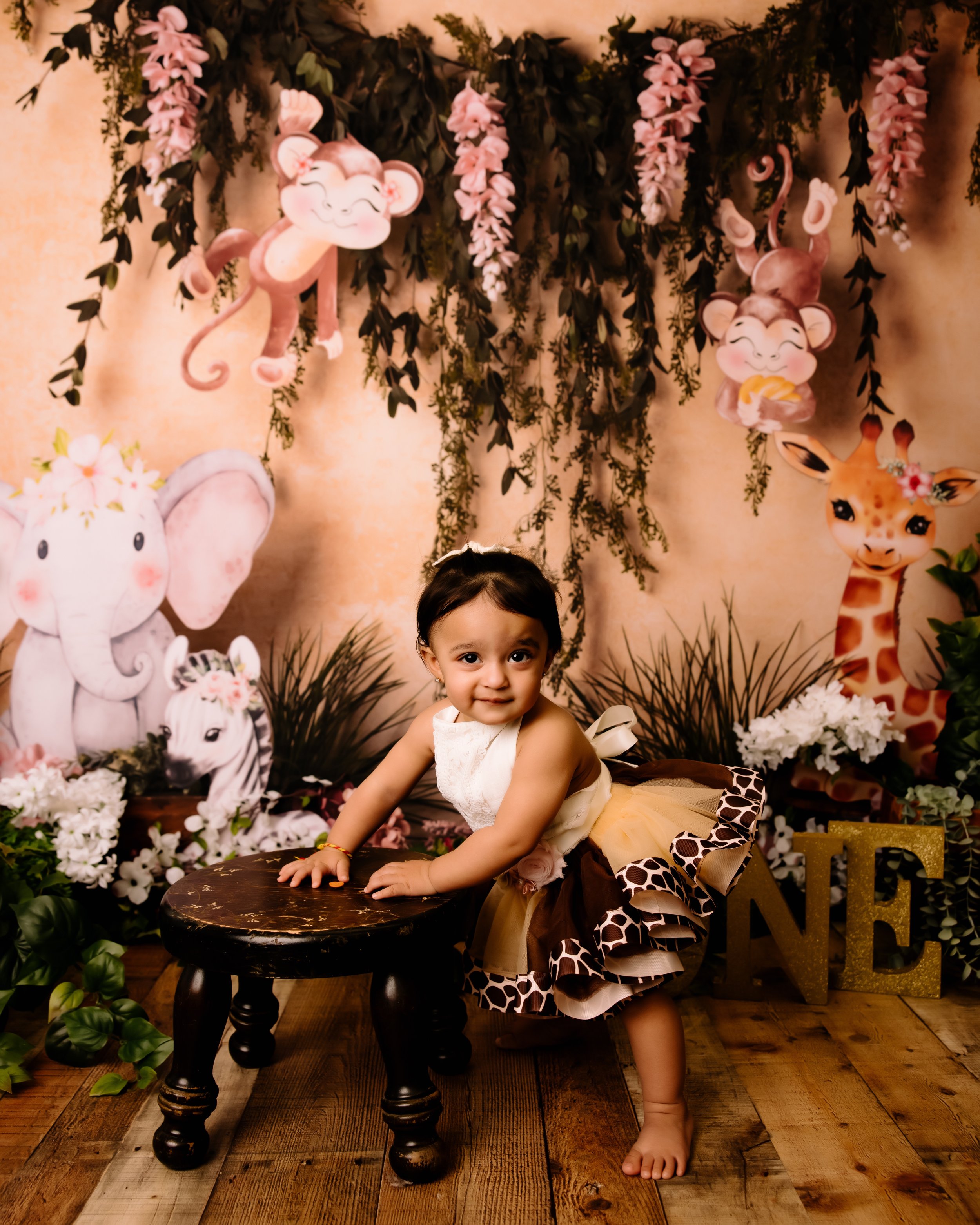 A young girl in a animal print and beige dress poses with her hands on a small dark wooden stool, in front of a jungle animal themed backdrop with pink flowers, hanging greenery, and illustrations of elephants, monkeys, giraffes, and zebras.