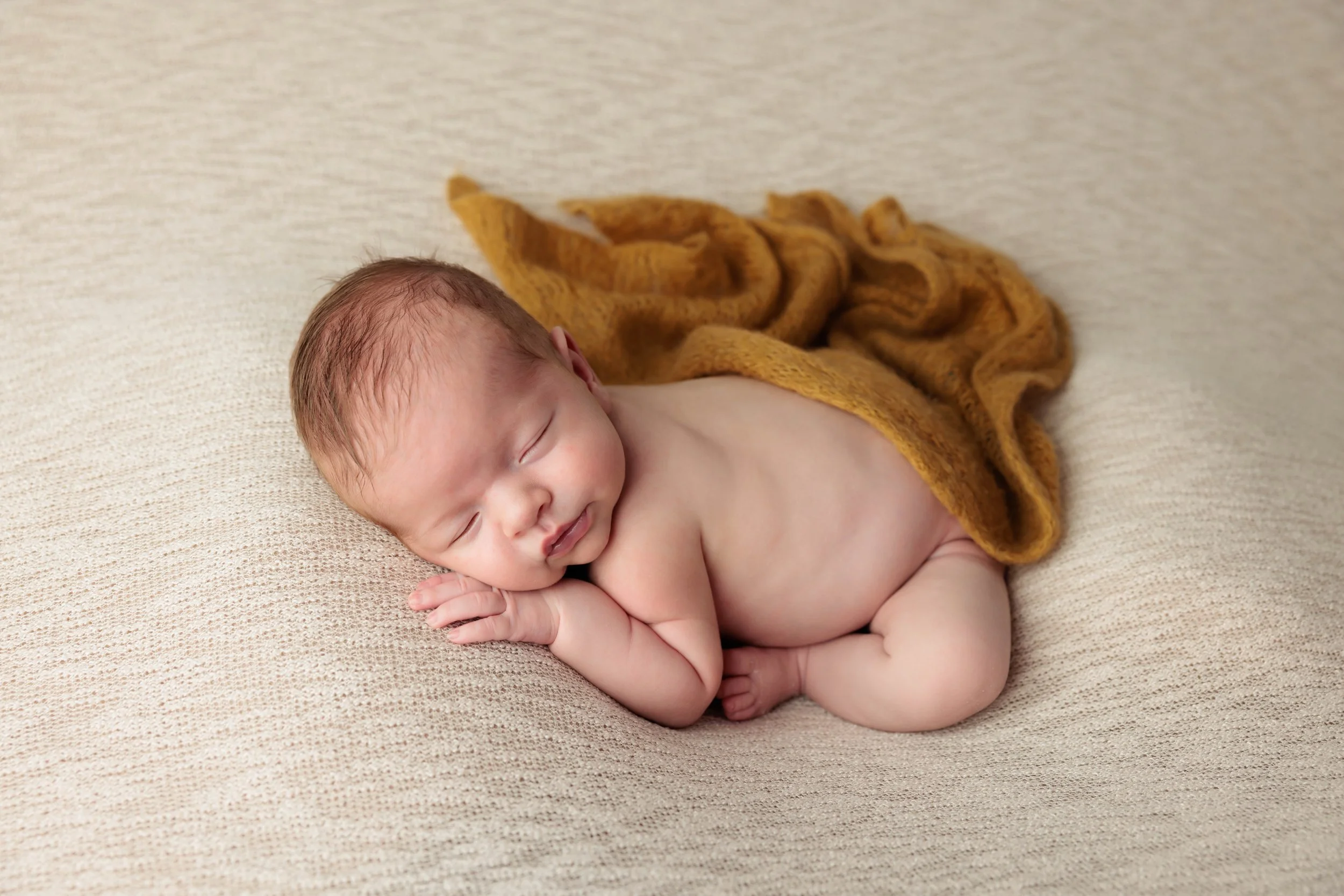 A sleeping newborn baby lying on a soft beige surface, partially covered with a mustard yellow blanket.