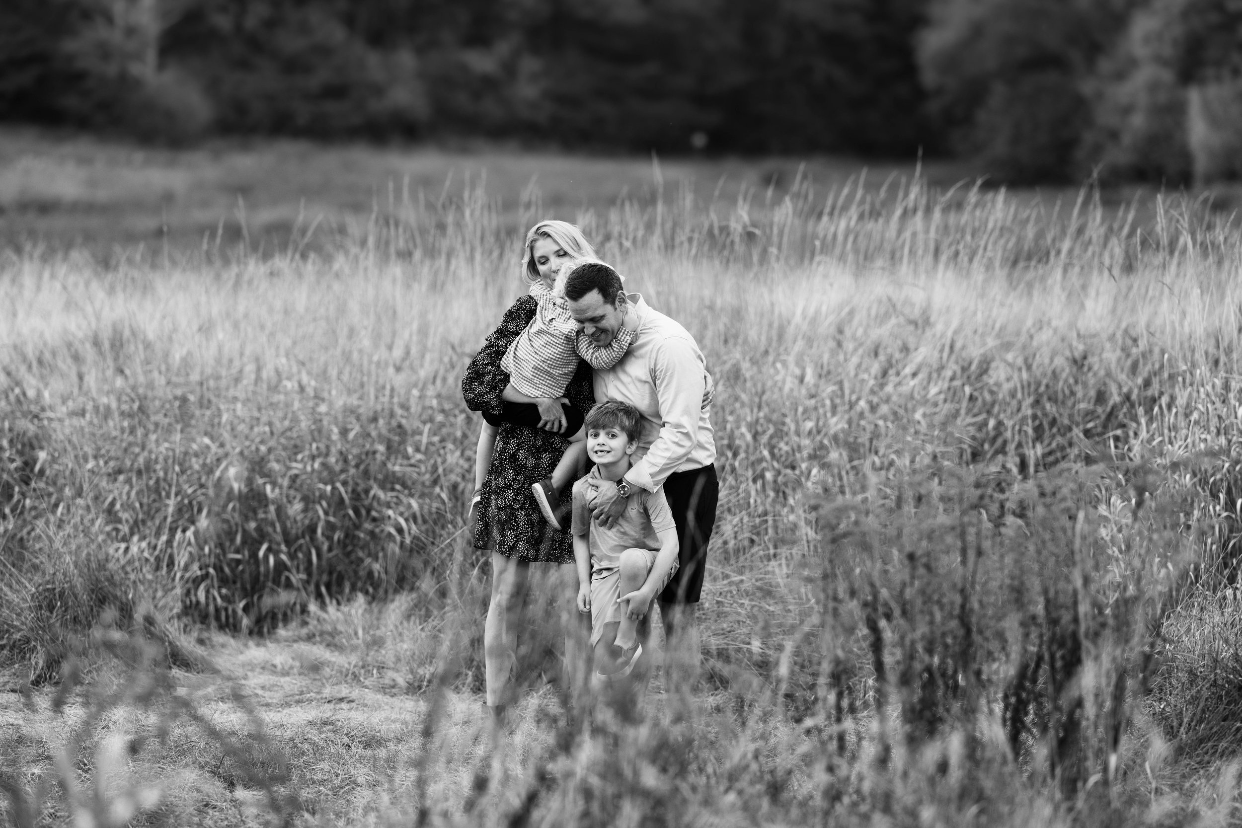 A black and white photo of a family of four in a grassy field: a woman, a man, and two children, smiling and embracing.